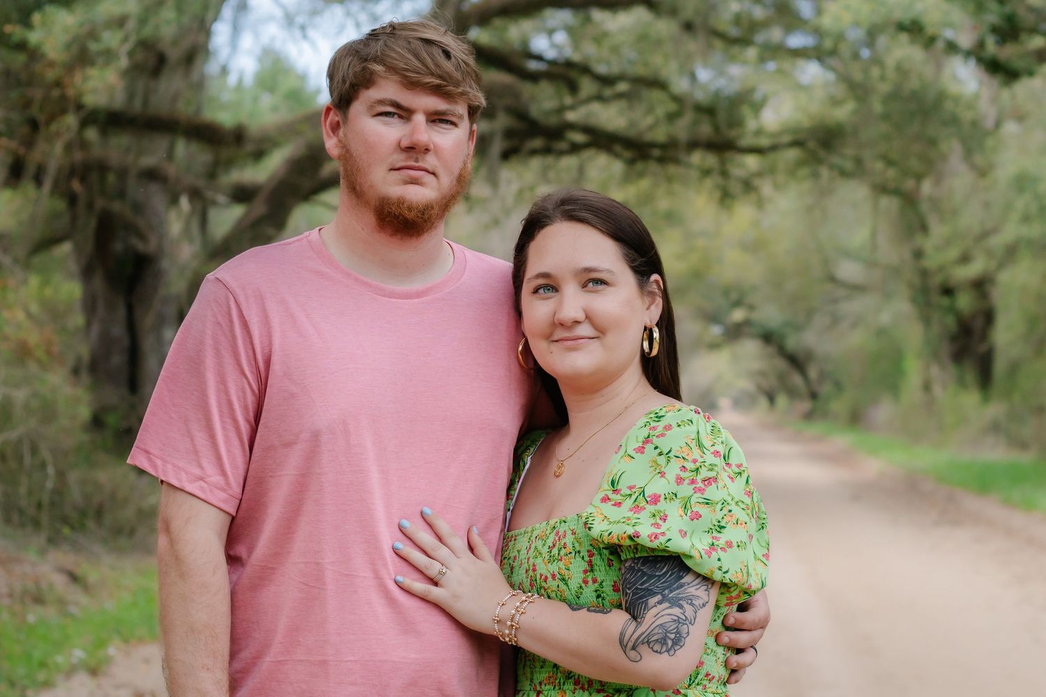 Couple embracing on a rustic dirt path framed by tall green trees in natural light.