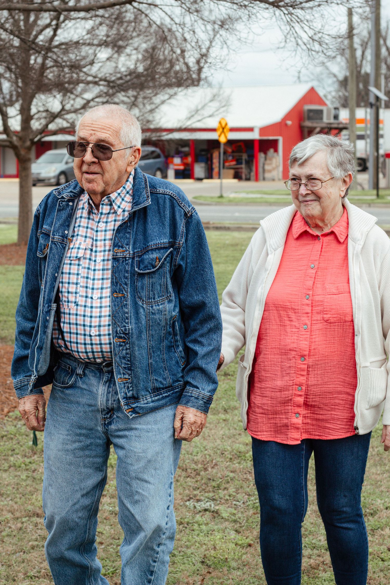 Senior couple walking together outdoors near a red building on a cloudy day.