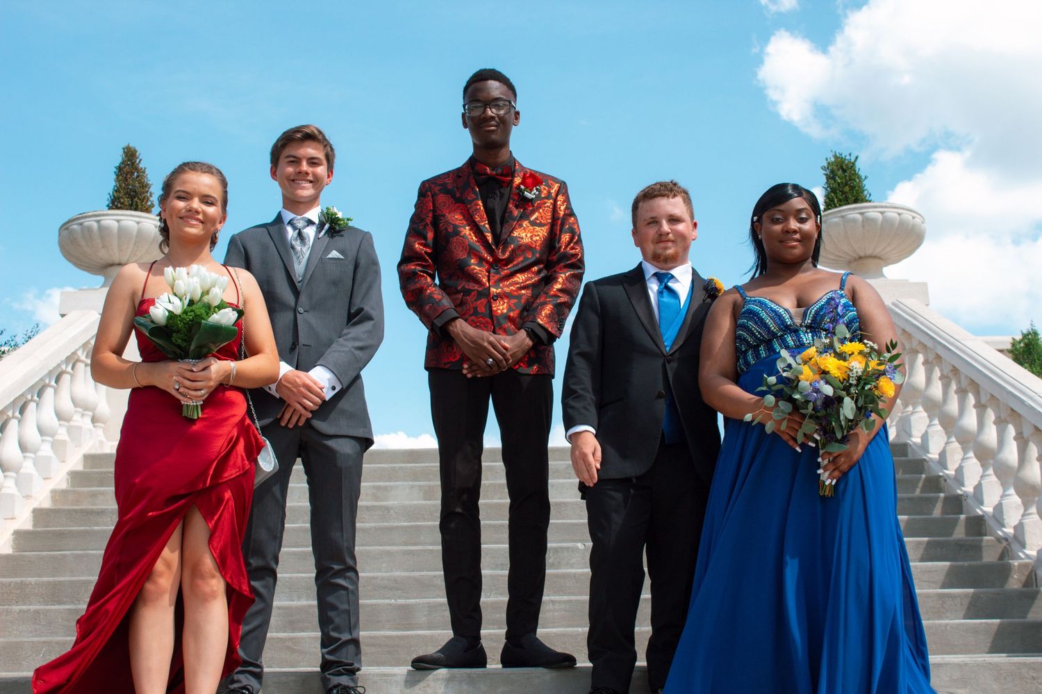 Group of five prom students posing on white marble stairs in formal attire with bouquets.