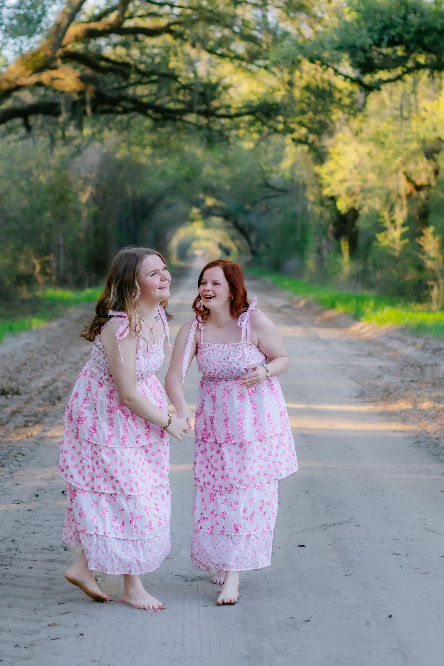 Two barefoot women in matching pink floral dresses laugh and walk together on a scenic dirt road.