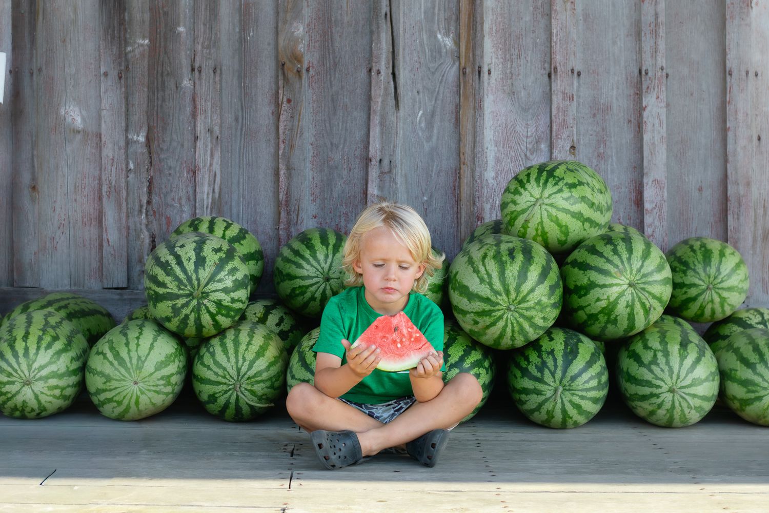 Young blonde boy in green shirt sits cross-legged eating watermelon slice surrounded by pile of whole watermelons outdoors.