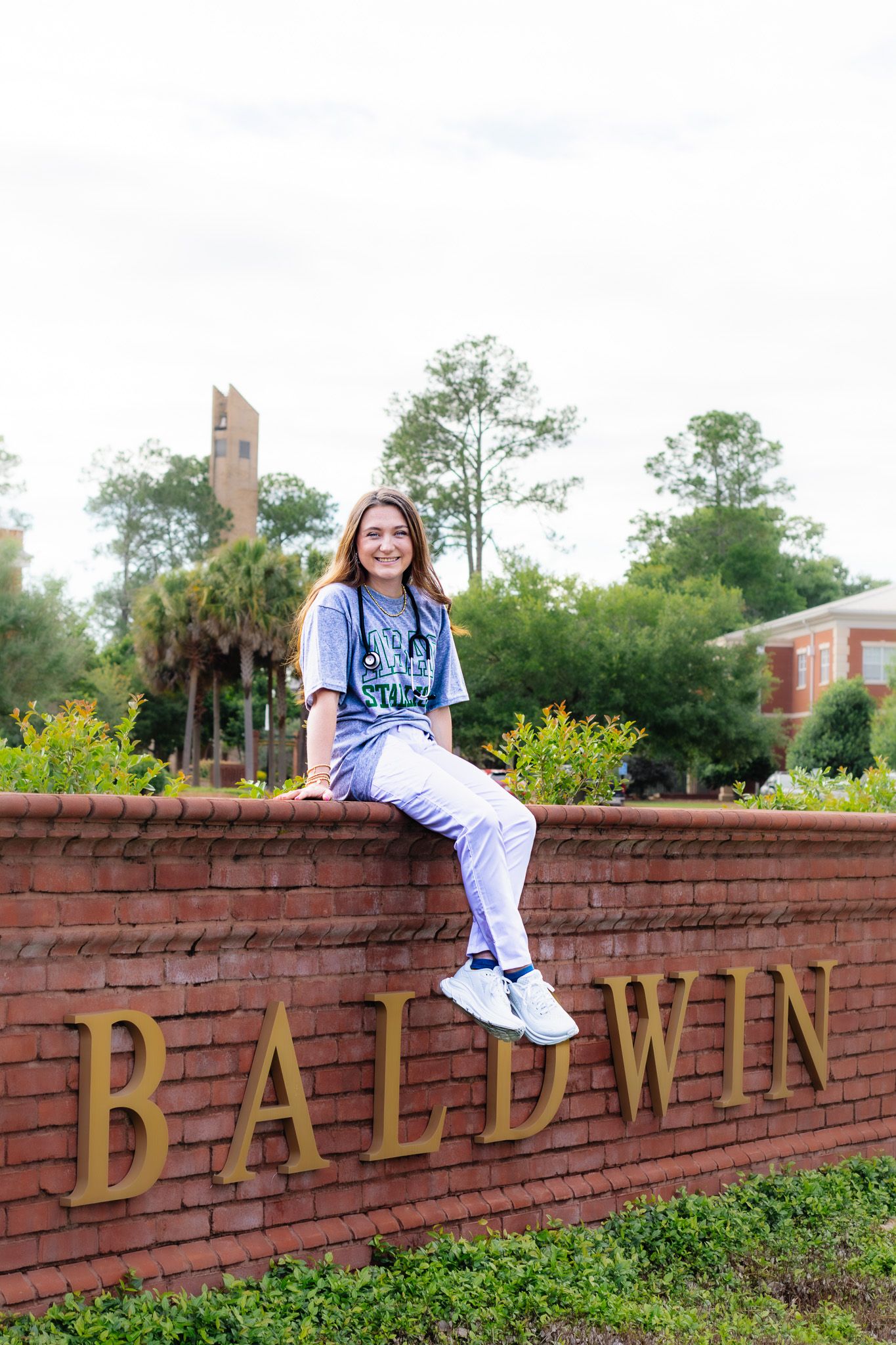 Smiling female student sits on Baldwin brick university entrance sign wearing college sweatshirt.