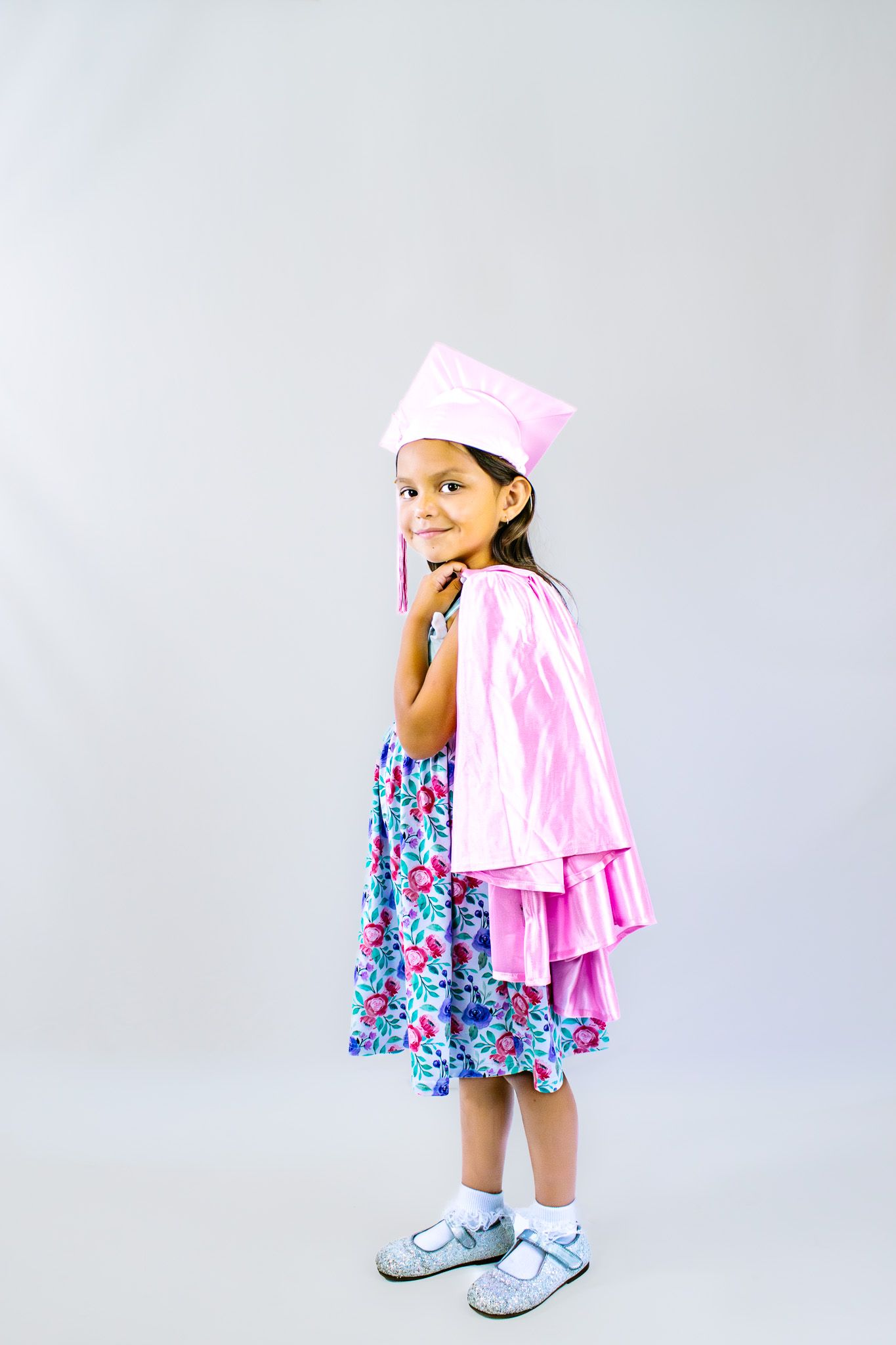 Young girl in pink graduation cap and gown over floral dress, smiling and posing against gray background.