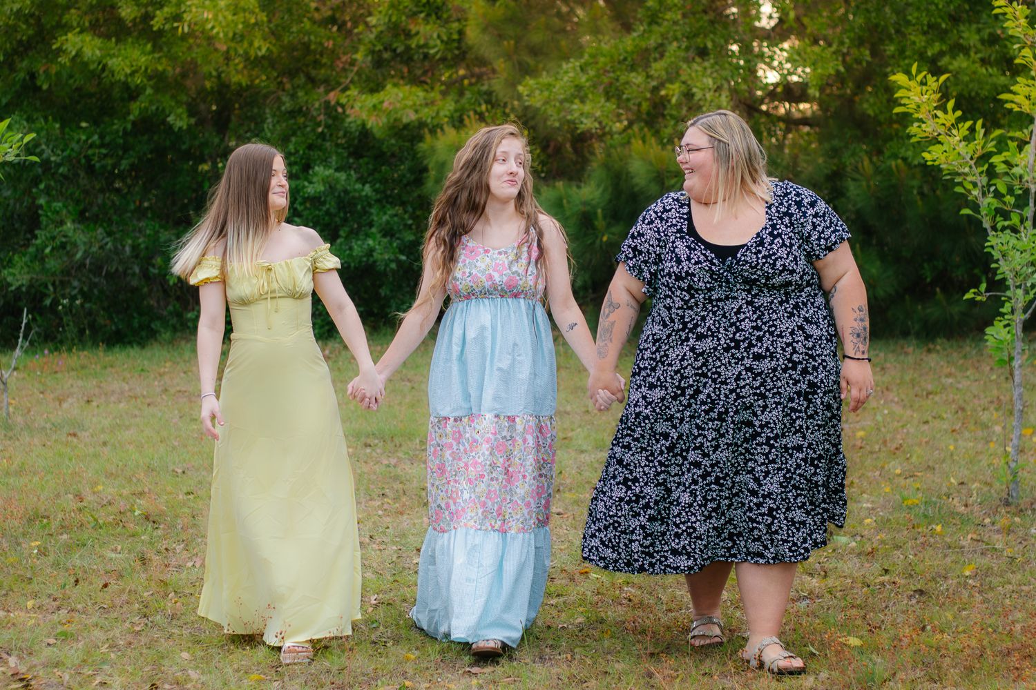 Three women in floral dresses holding hands and walking together outdoors in a lush green park setting.