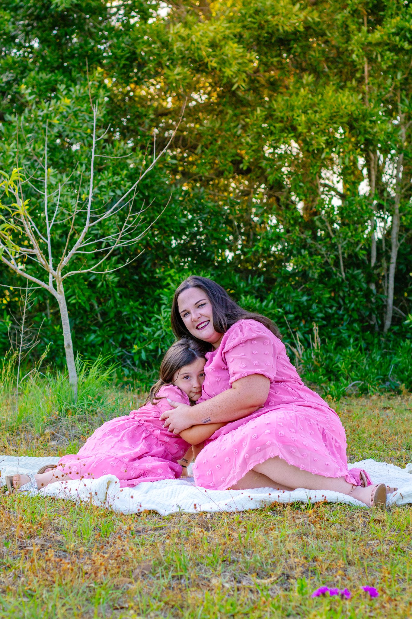 A woman and young girl in matching pink dresses sit together on a blanket outdoors, smiling and hugging.