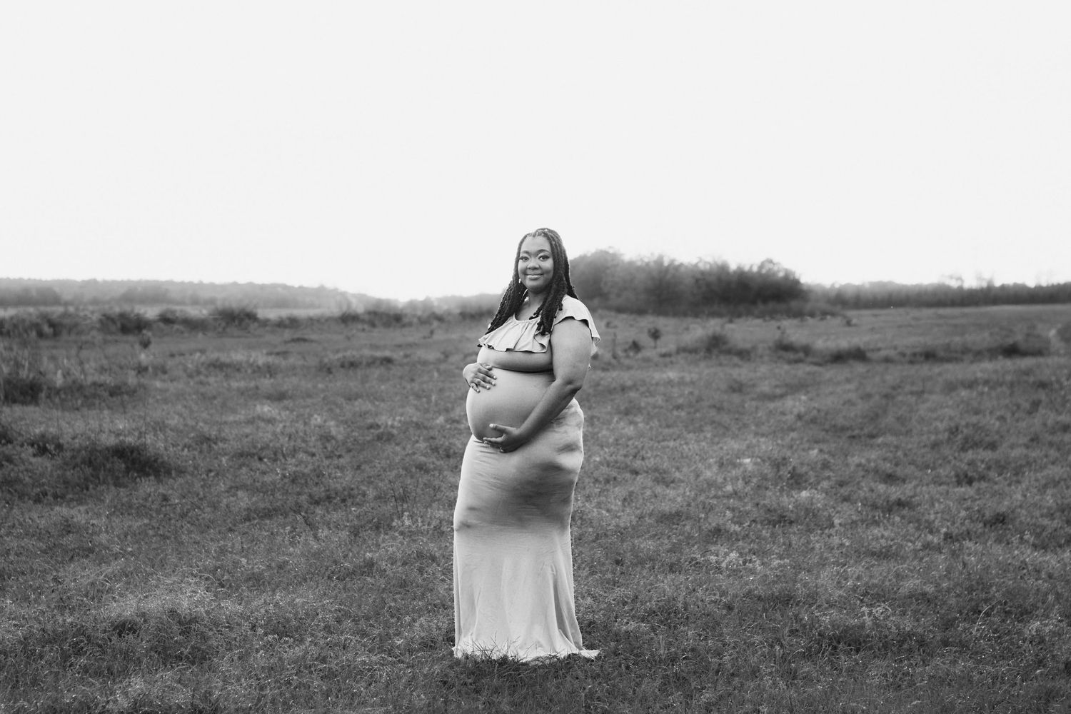 Smiling pregnant woman in white gown cradles baby bump in open field in black and white photo.