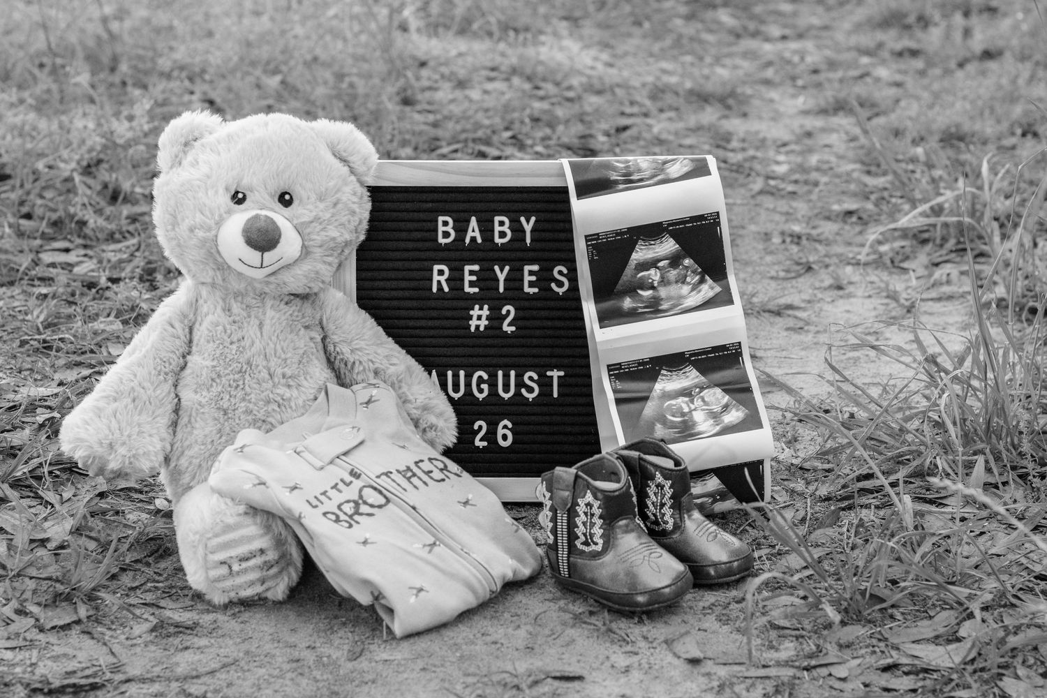 Black and white flat lay of Baby Reyes #2 announcement with teddy bear, ultrasound photos, and baby shoes.