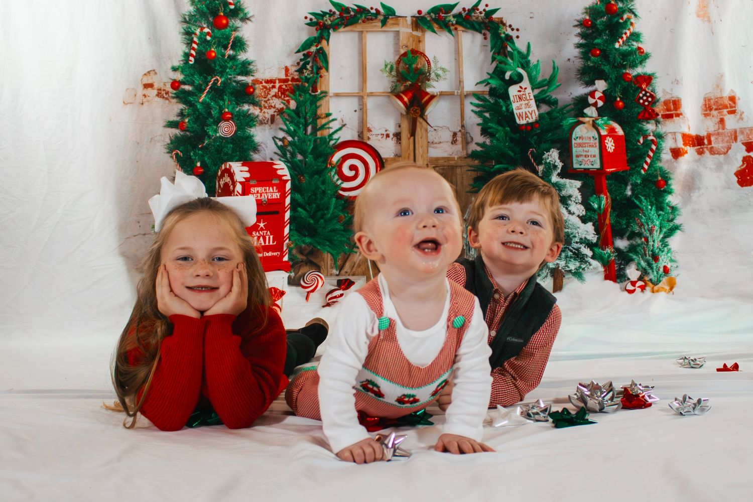 Three children pose together in front of festive Christmas holiday backdrop indoors.