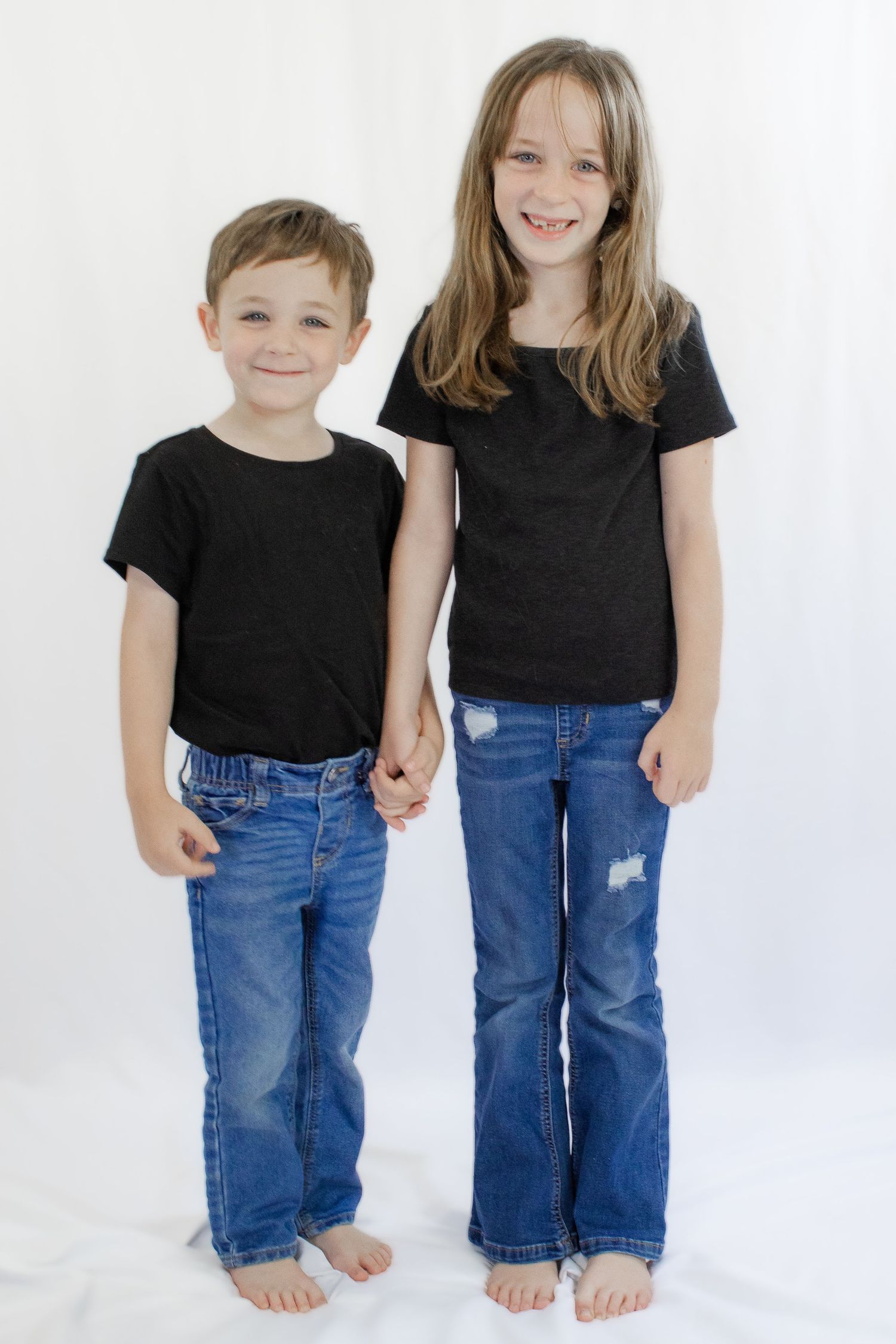 Two barefoot children in matching black t-shirts and blue jeans holding hands and smiling against a white background.