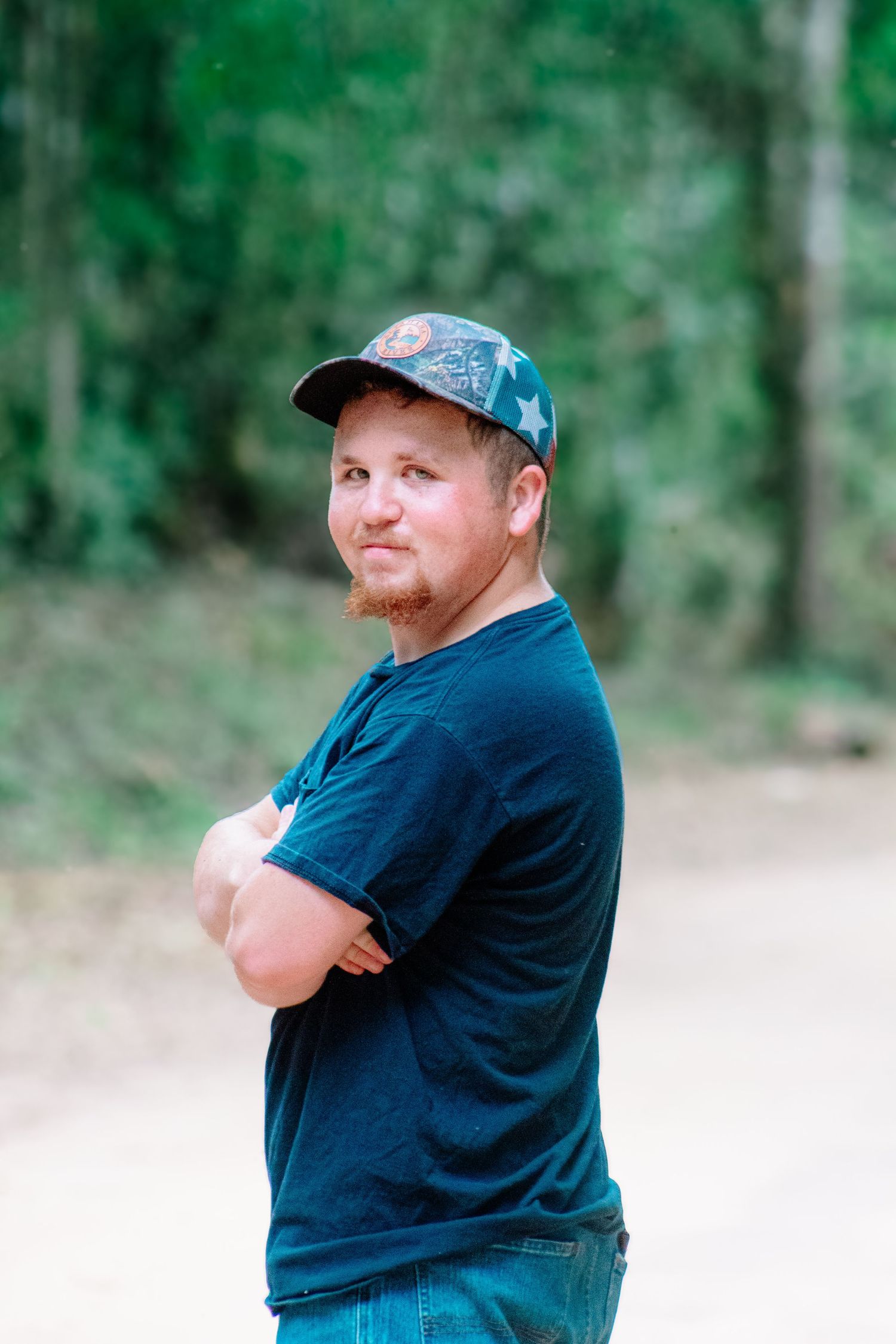 Man in black t-shirt and camo cap standing with arms crossed on a wooded dirt road.