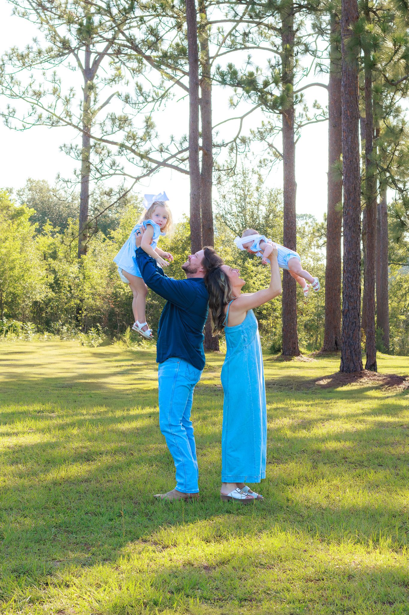 Joyful couple lifts two young children in the air outdoors, wearing coordinating blue outfits among tall pine trees.