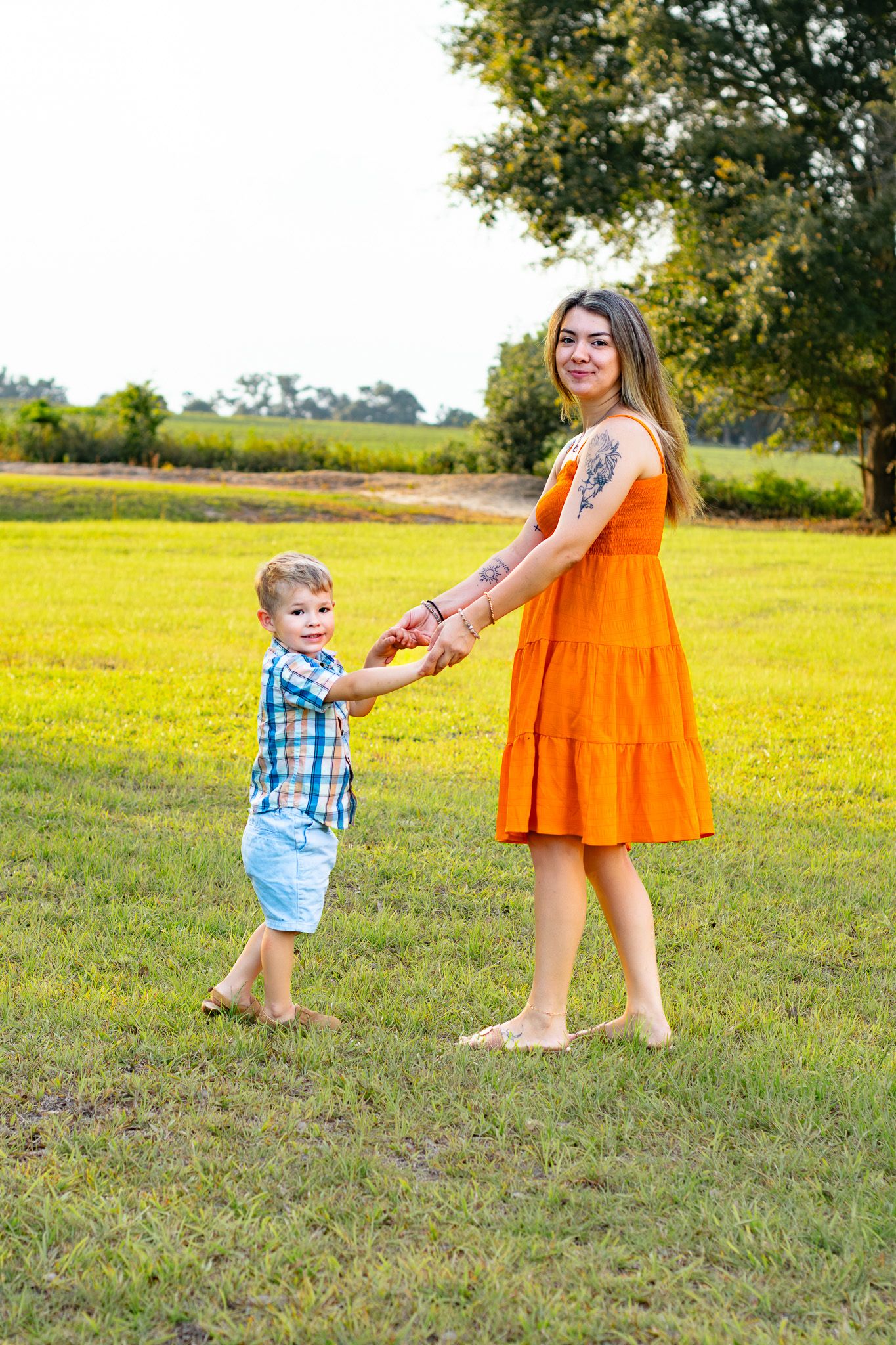 A mother in an orange dress and her young son in a plaid shirt hold hands barefoot in a sunny meadow.