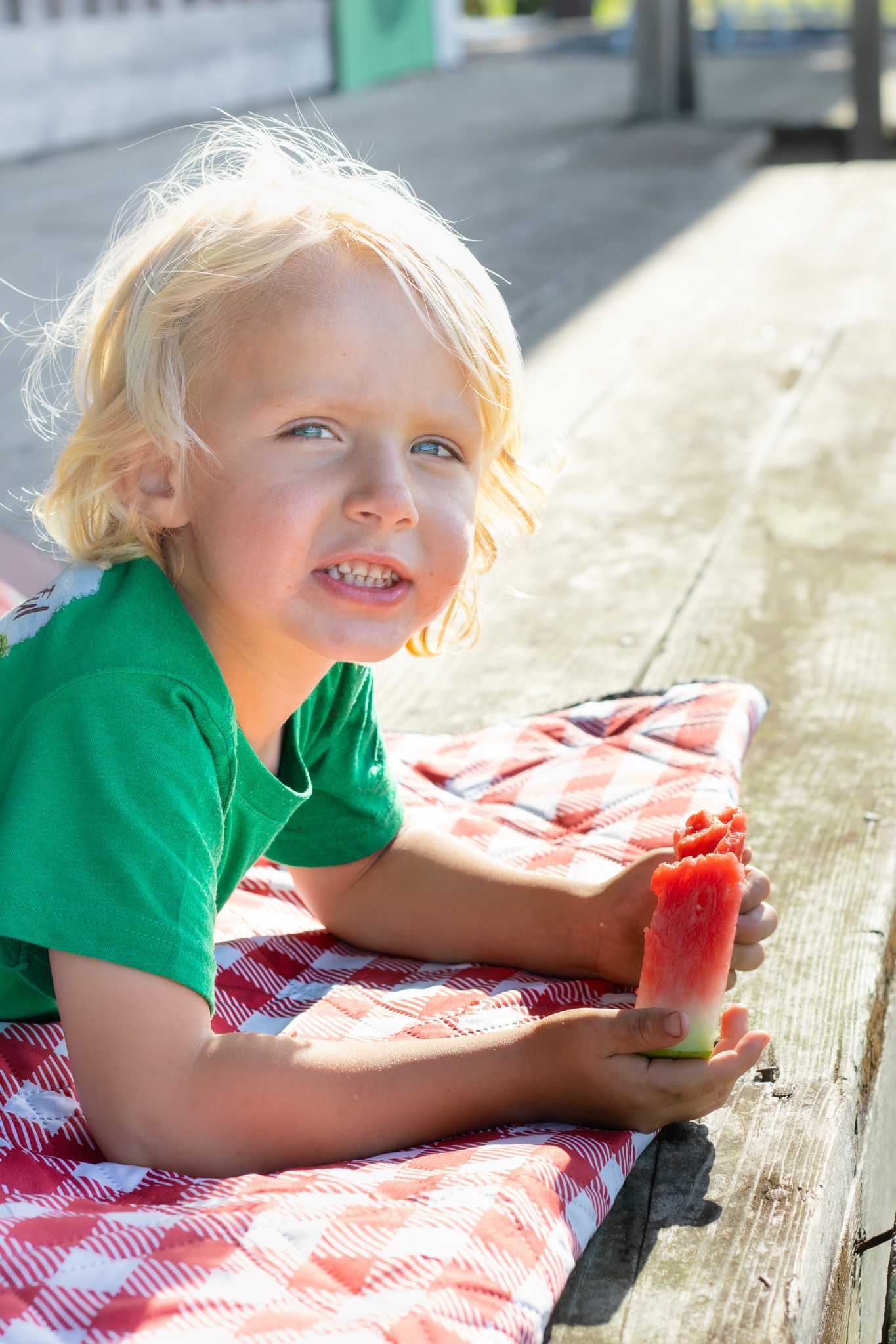 Blonde toddler in green shirt eating watermelon slice while lying on red checkered blanket on wooden deck.