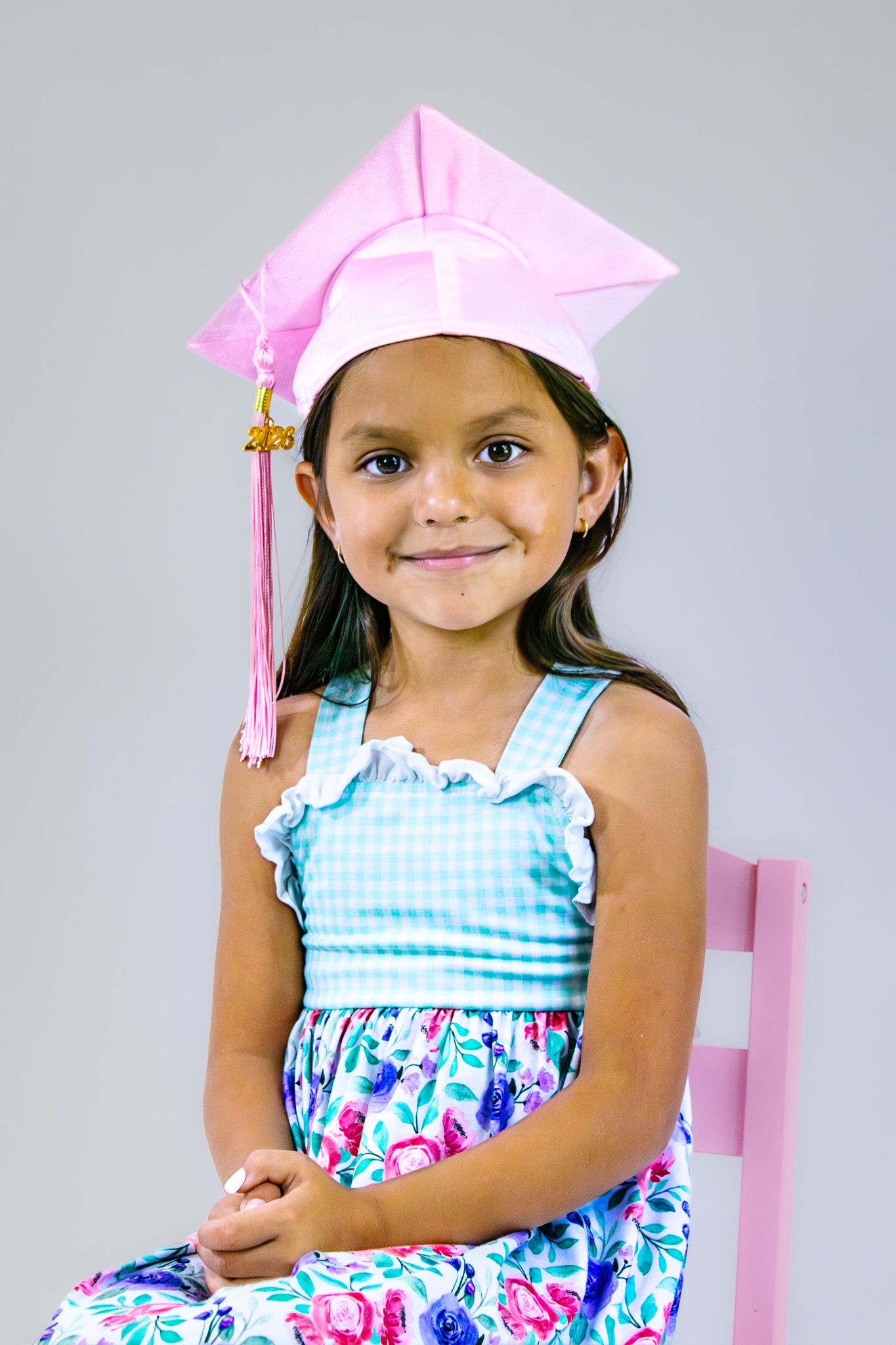 A young girl wearing a pink graduation cap and floral dress sits smiling on a pink chair for a preschool graduation photo.