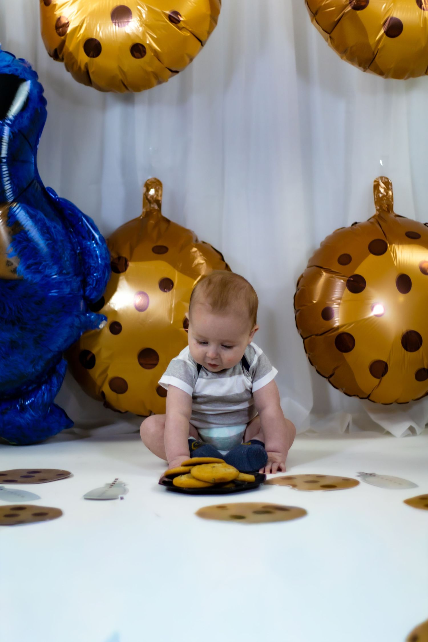 Baby boy in white onesie sitting among Cookie Monster themed balloons and chocolate chip cookie props.
