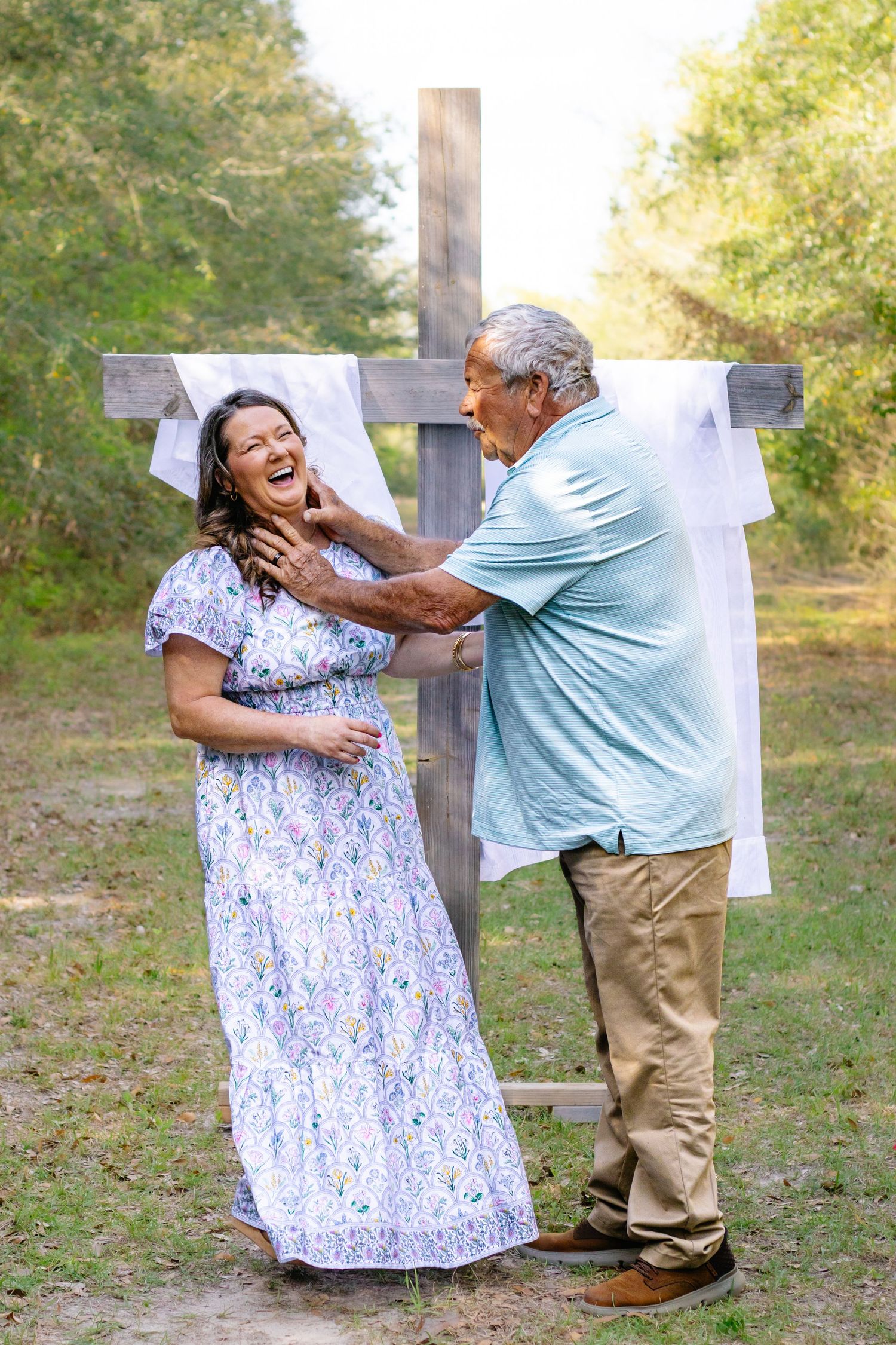Couple laughing joyfully in front of a wooden cross, man in blue polo gently touching womans face.
