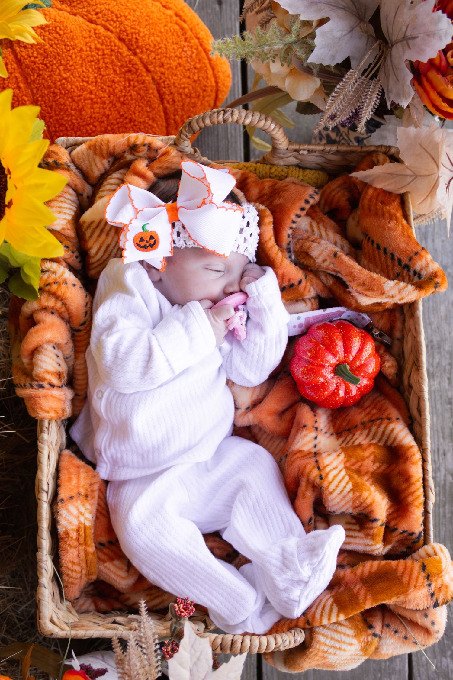 A newborn baby sleeps peacefully in a wicker basket surrounded by fall pumpkins and sunflowers.