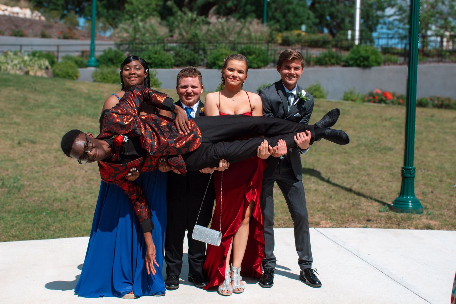 Four teenagers in prom attire playfully posing outdoors, with one person being carried horizontally.