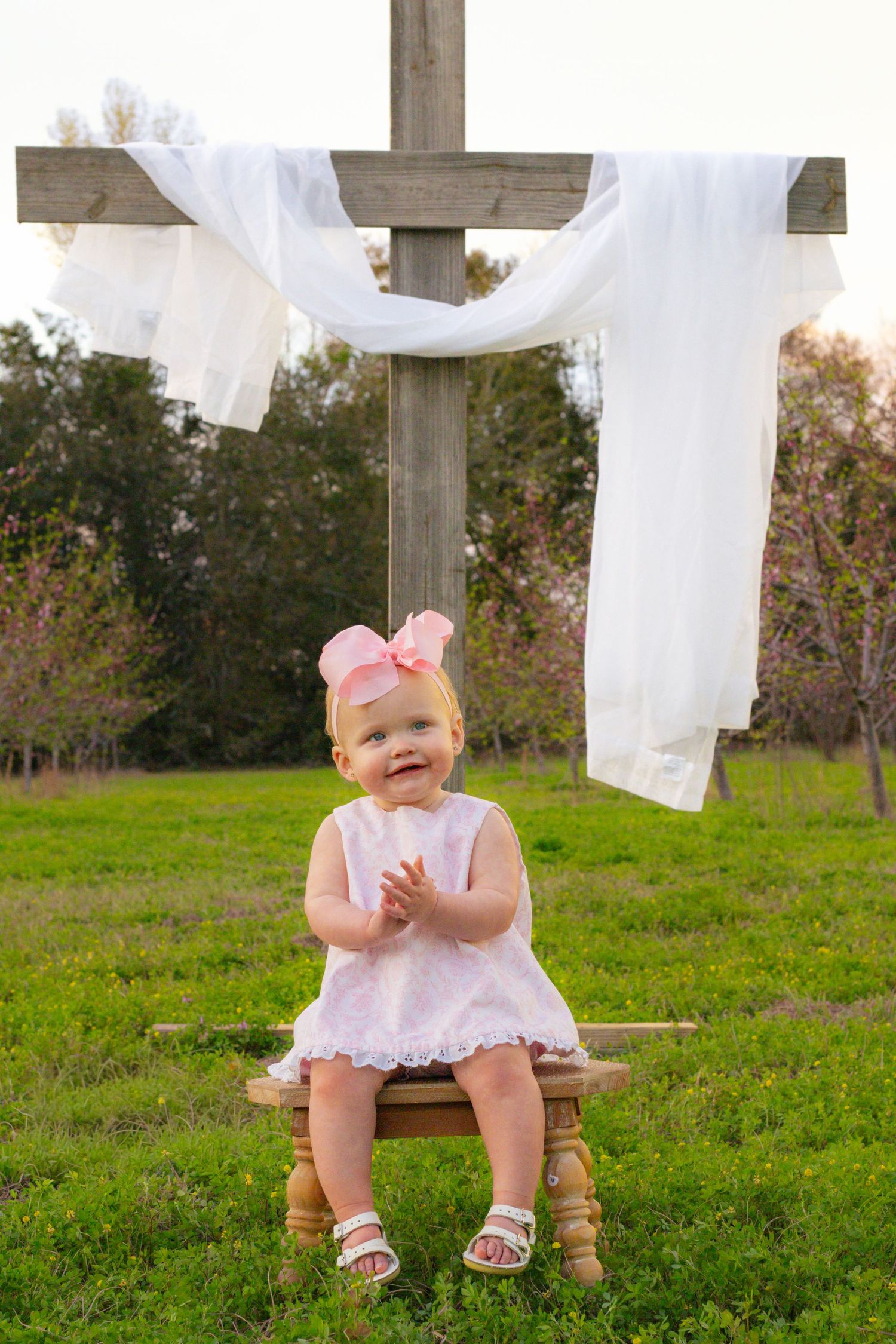 Smiling baby girl in white dress with pink bow sits on wooden stool in front of a rustic outdoor cross.