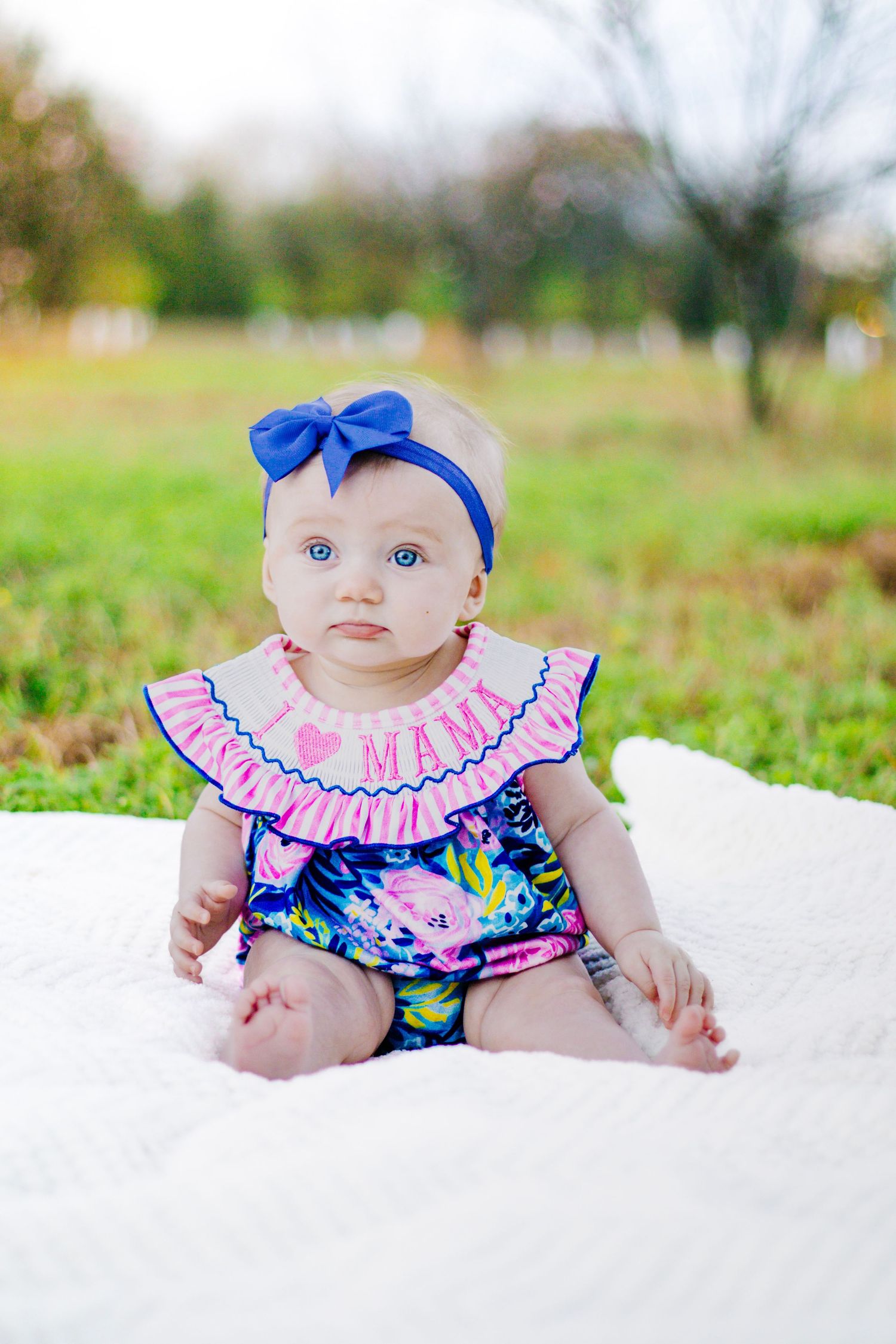 Baby girl in floral romper with blue bow headband sitting on white blanket outdoors.