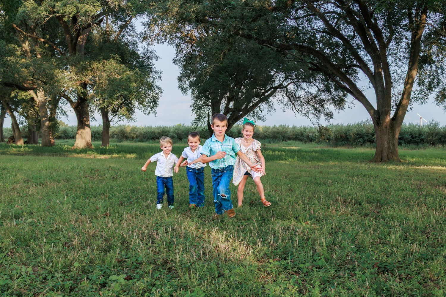 Four young children running and playing together in a lush green park surrounded by tall trees.