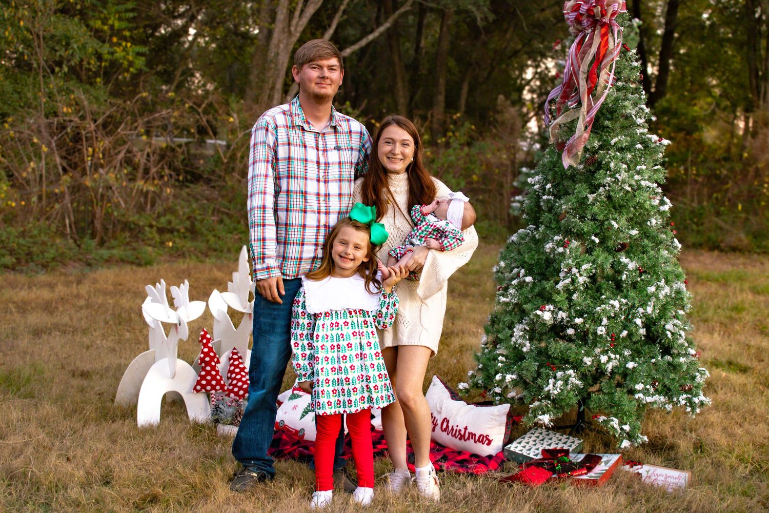 Family of four posing beside decorated Christmas tree outdoors with white reindeer decorations.