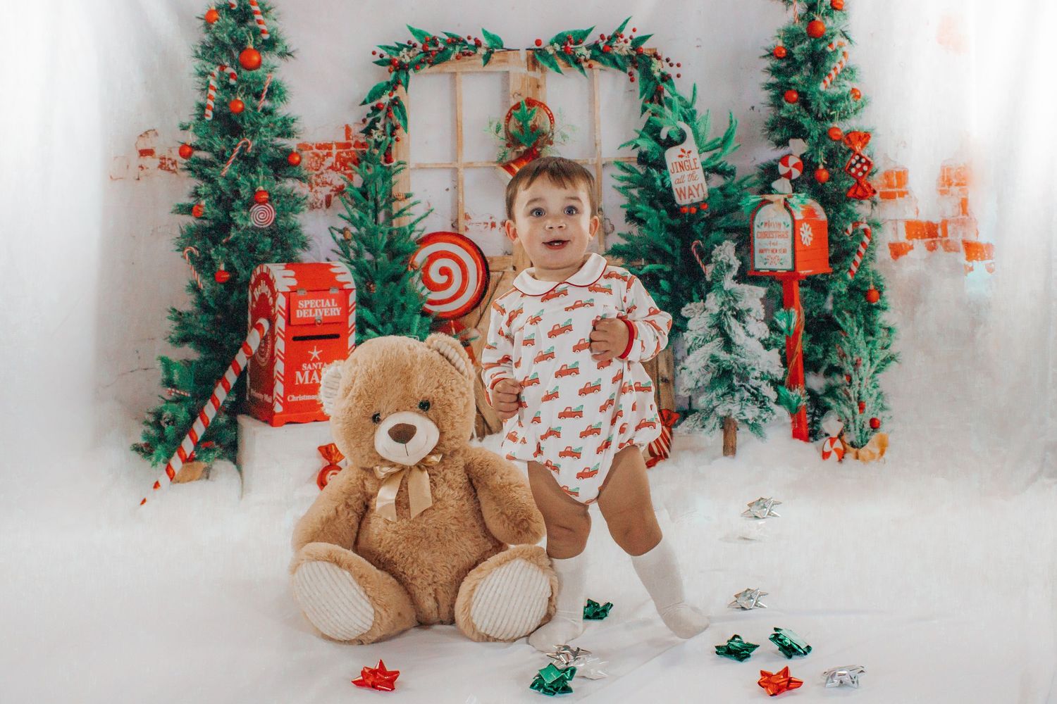 Baby in Christmas onesie stands beside teddy bear in front of holiday backdrop.