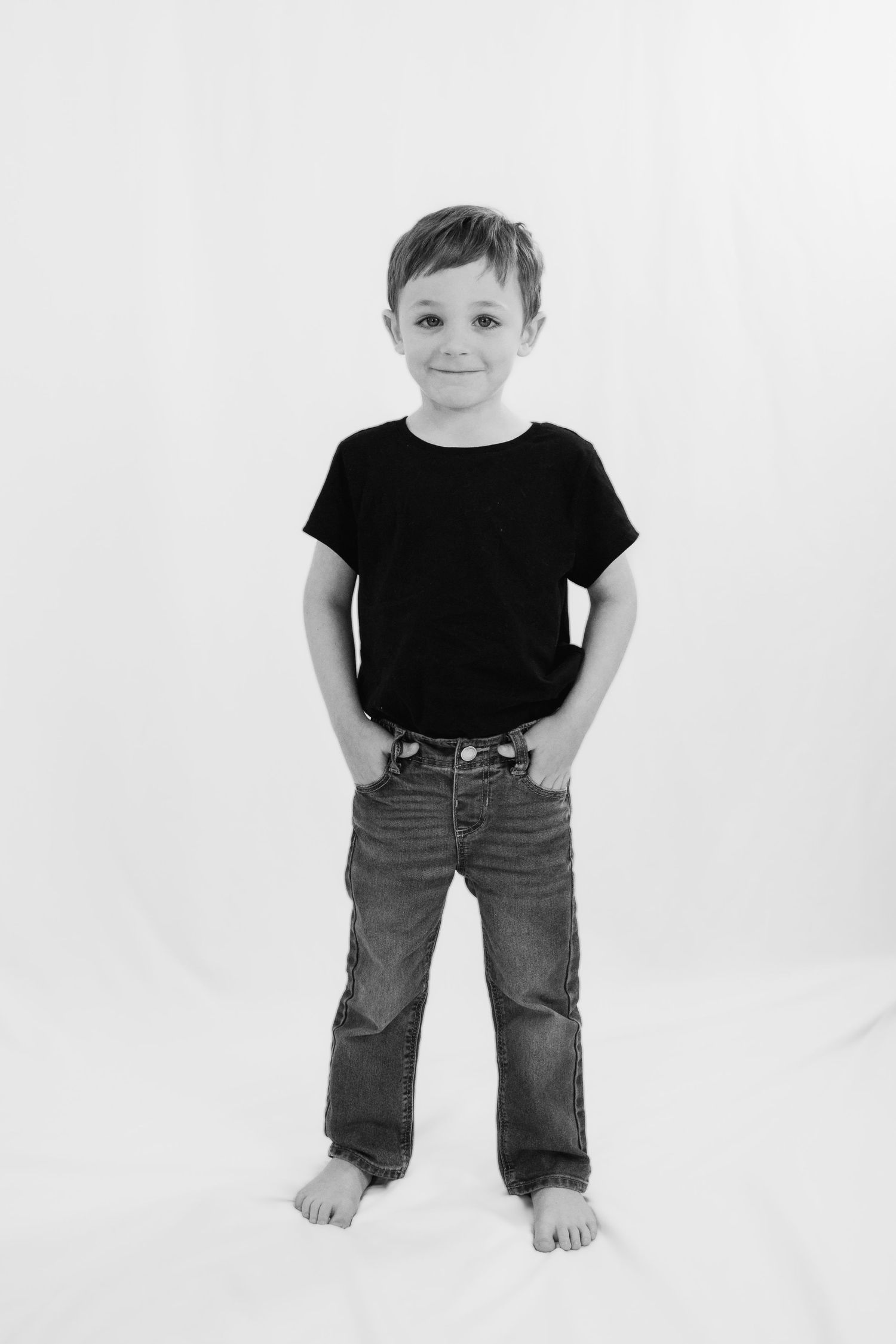 Black and white portrait of a young boy in a black t-shirt and jeans standing barefoot against a white background.