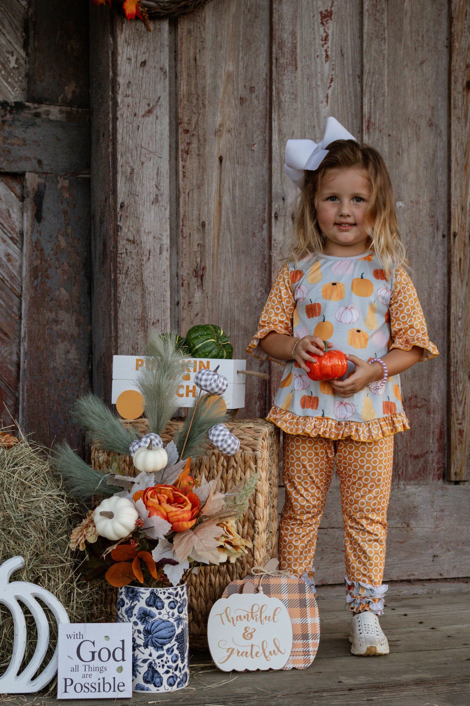 Young girl in fall pumpkin outfit holding small pumpkin beside harvest decorations and barn wood backdrop.
