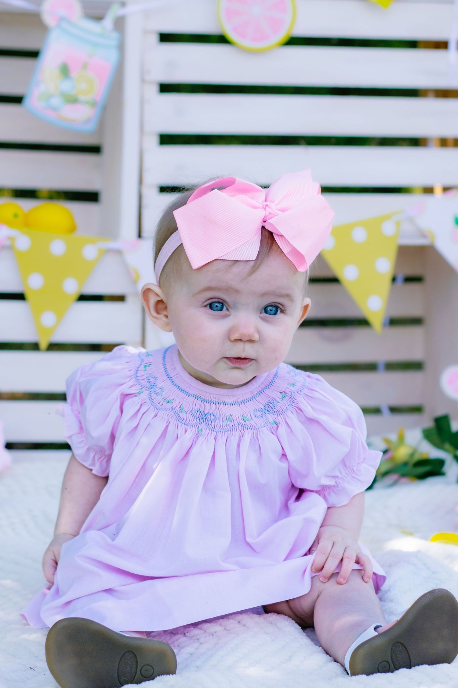Cute baby girl with blue eyes in pink smocked dress and large pink bow headband.
