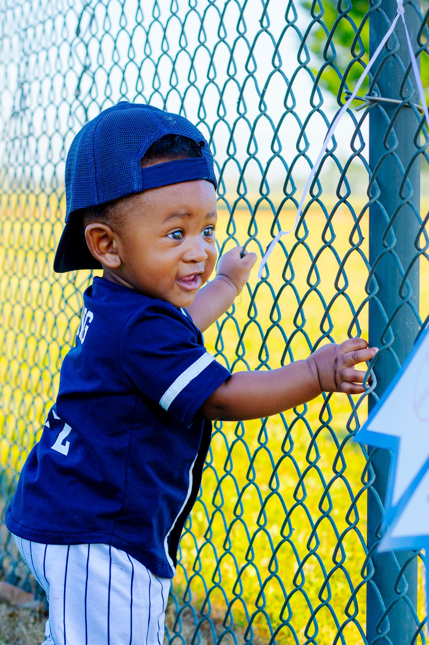 Young boy in blue baseball uniform and cap smiles while holding a green chain-link fence outdoors.
