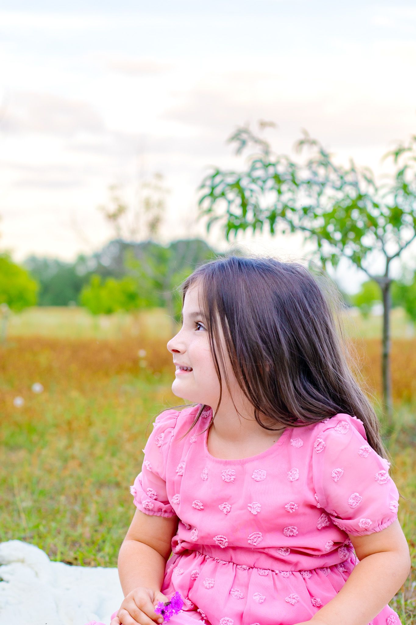 A young girl in a pink dress sits outdoors in a green field, smiling and looking to the side on a cloudy day.