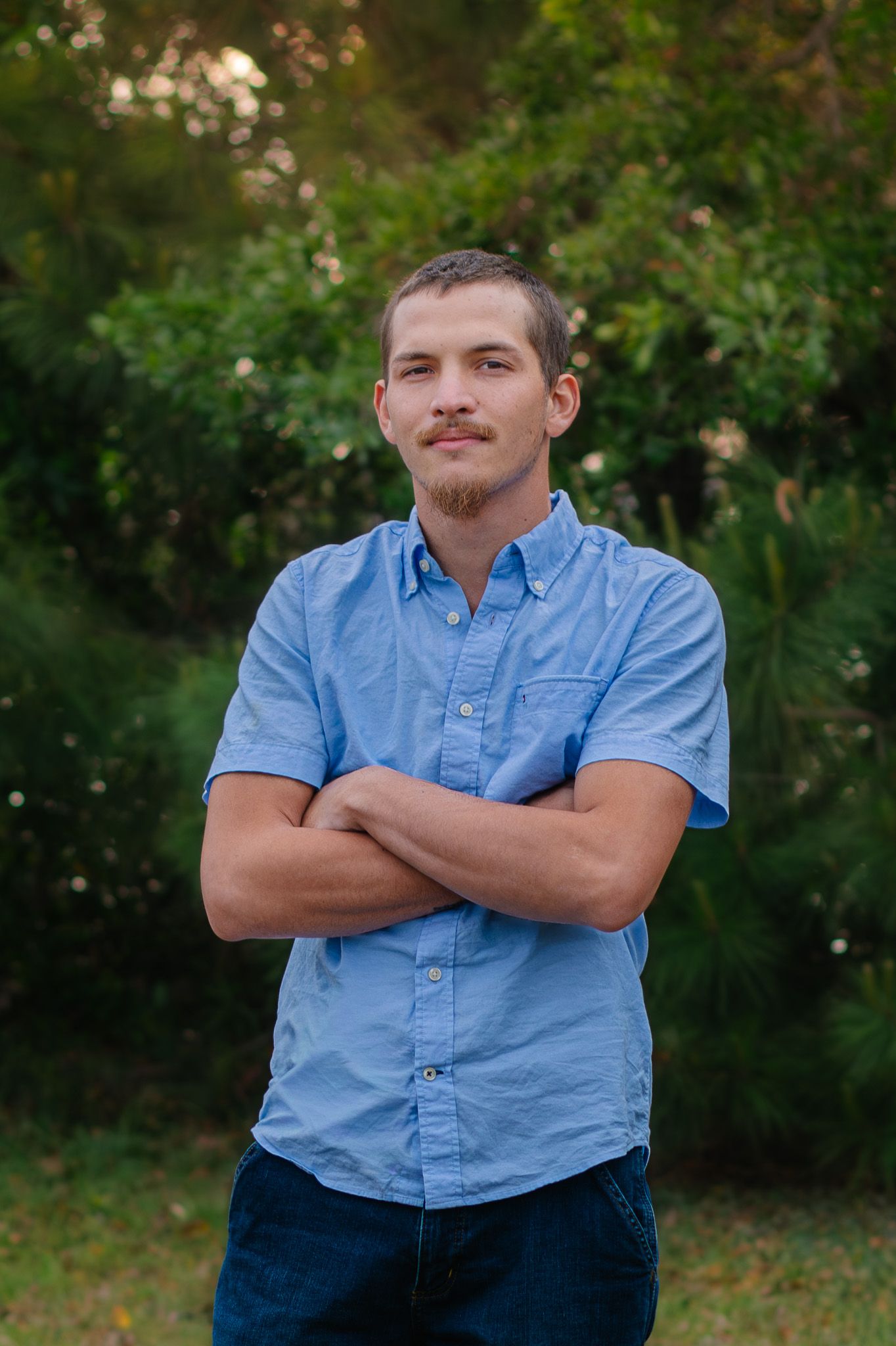 Young man with mustache in blue shirt standing outdoors with arms crossed smiling.