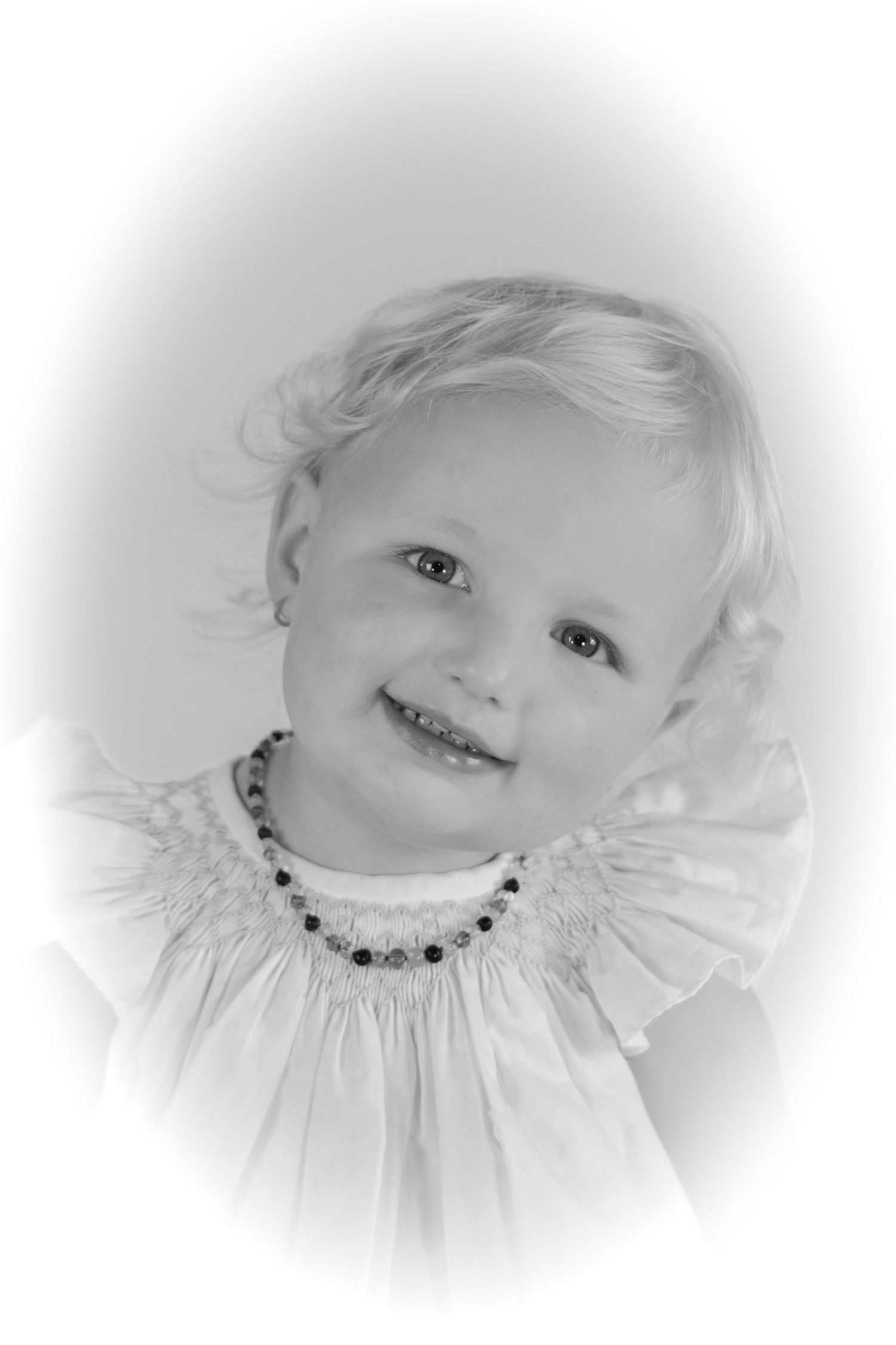 Black and white portrait of a smiling toddler girl wearing a ruffled dress and beaded necklace.