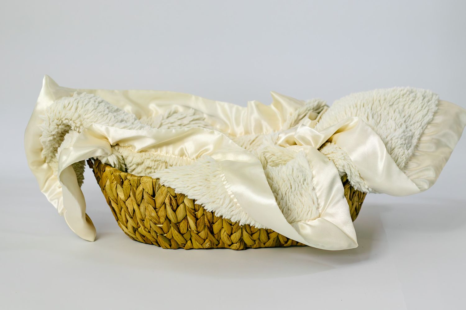 A woven straw basket lined with cream satin and soft white blanket on a gray background.