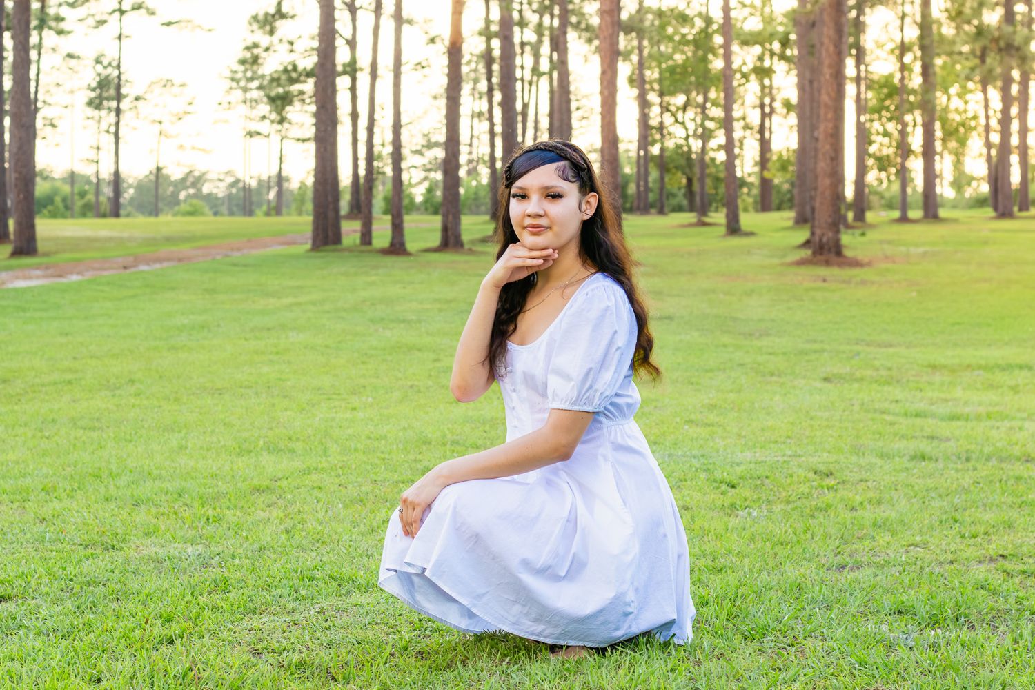 Young woman in white dress sitting gracefully on green grass among tall pine trees in golden hour light.