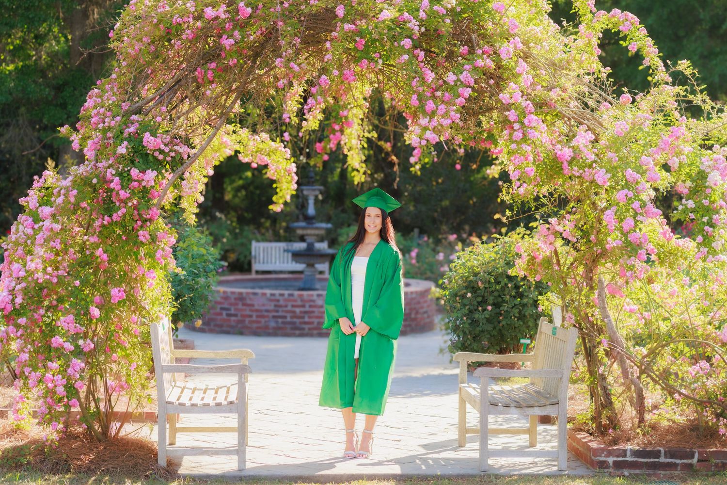 Graduate in green cap and gown stands under floral arch with pink blooms in a garden setting.