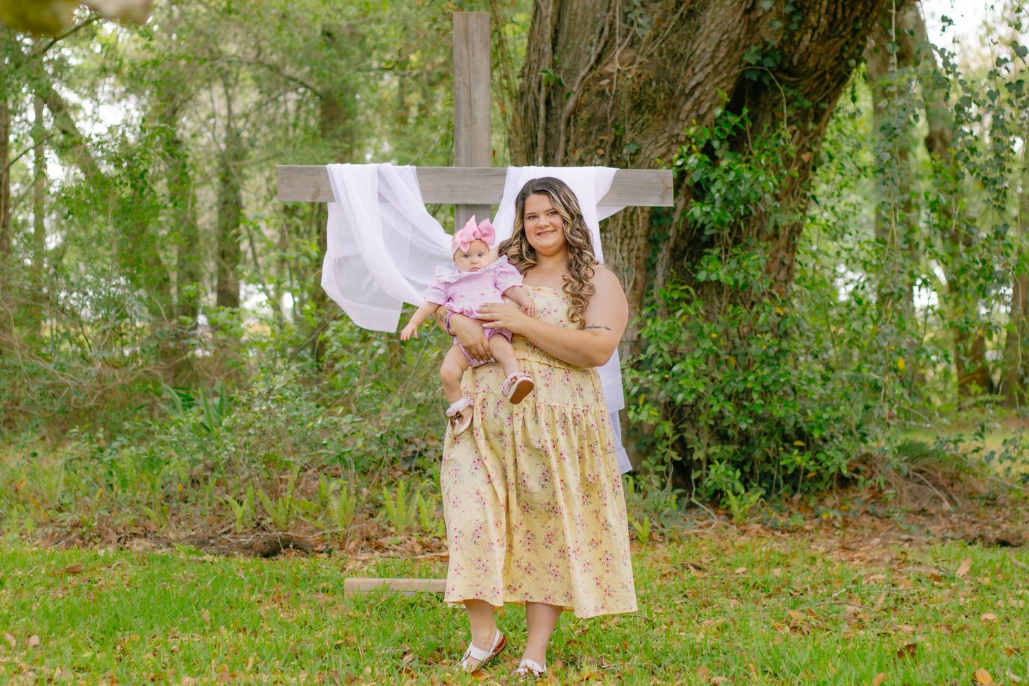 Mother in floral dress holding baby girl in pink outfit standing before a wooden cross outdoors.