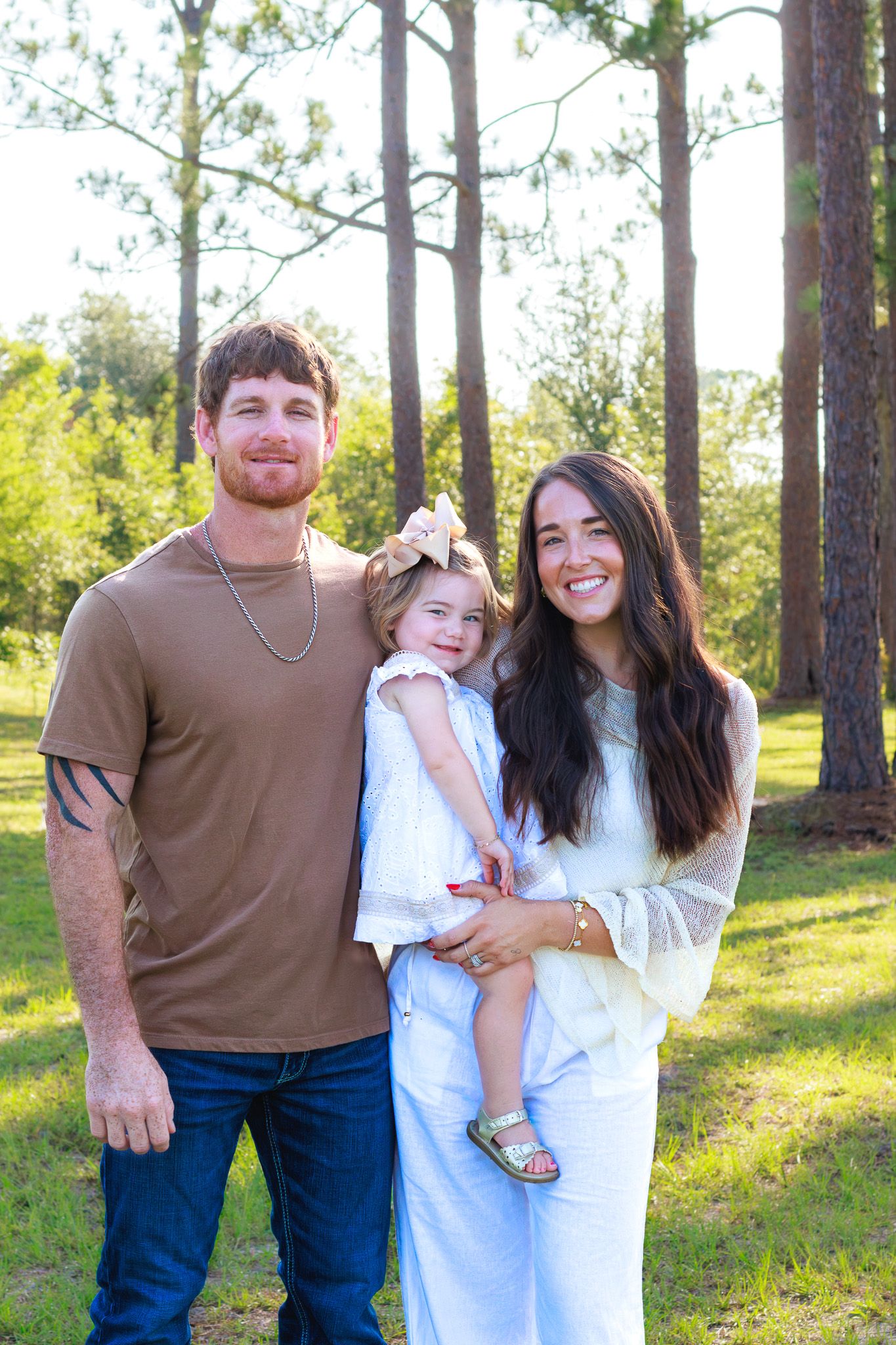 A smiling family of three poses outdoors among tall pine trees, with parents holding their young daughter in a white dress.