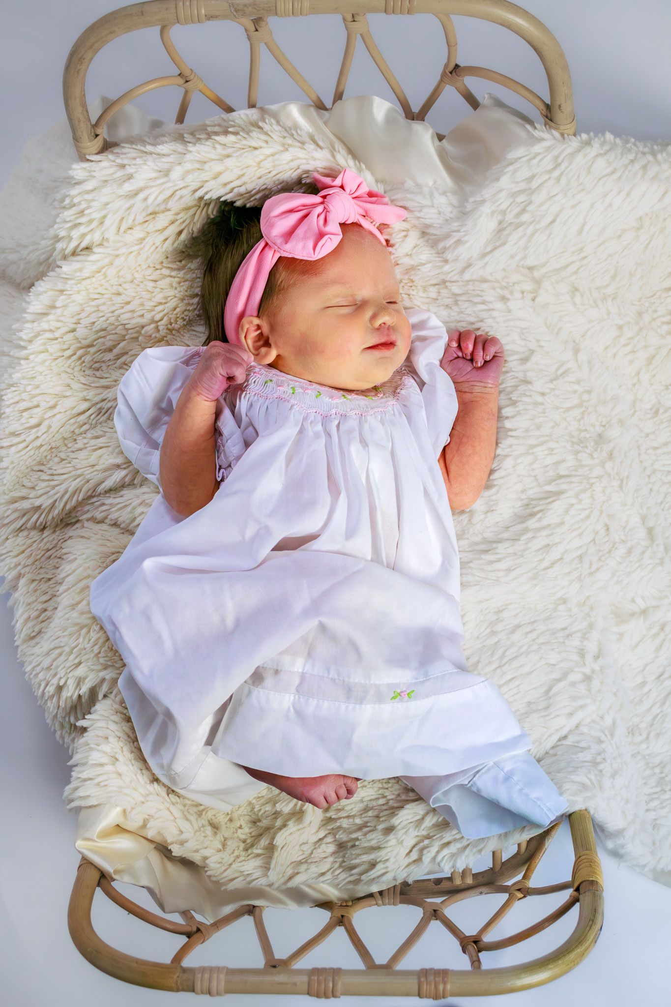 Newborn baby girl sleeping in a wicker bassinet wearing a white dress and pink bow headband.