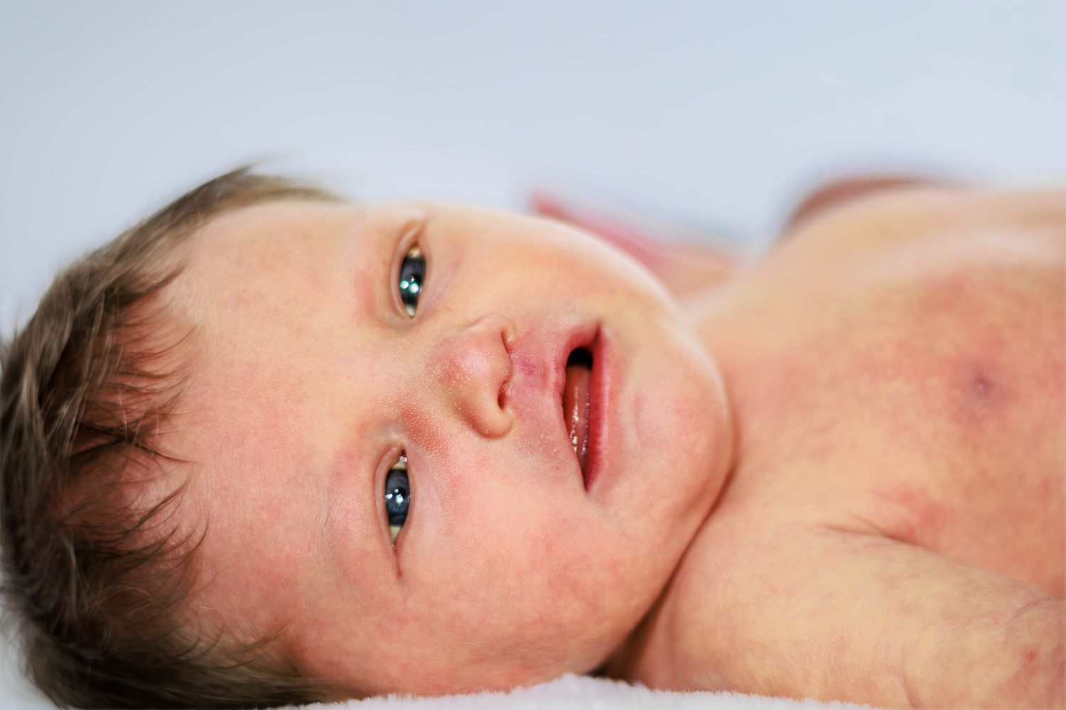 Close-up portrait of a newborn baby with blue eyes and rosy skin lying on a white blanket.