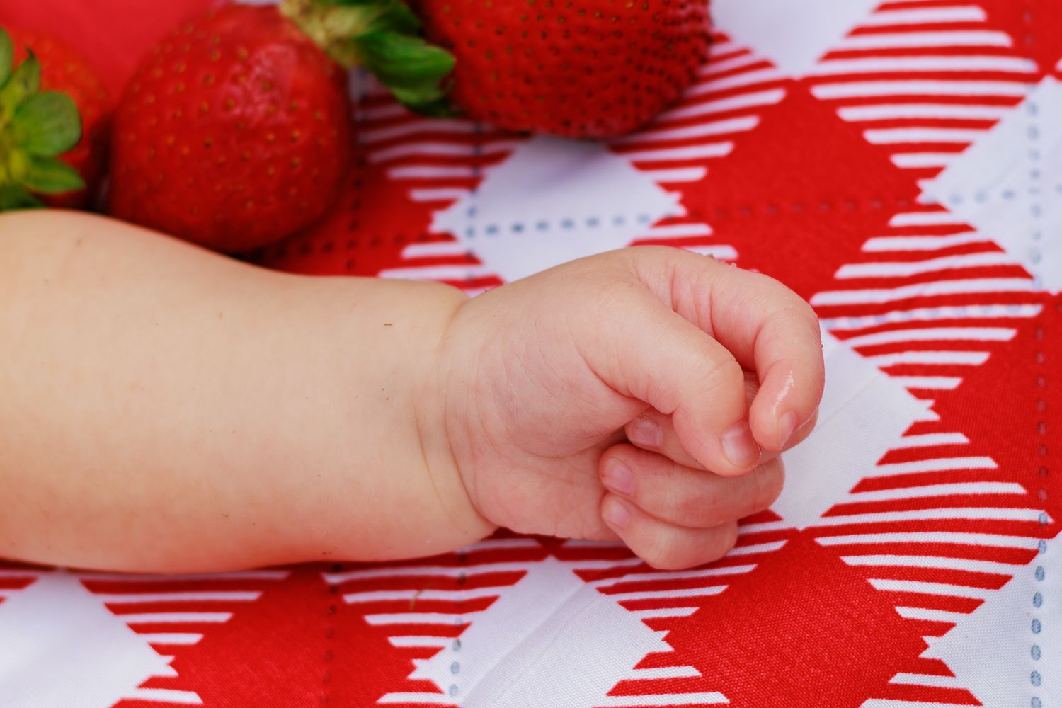 Close-up of a tiny baby hand resting on a red and white checkered blanket beside fresh strawberries.