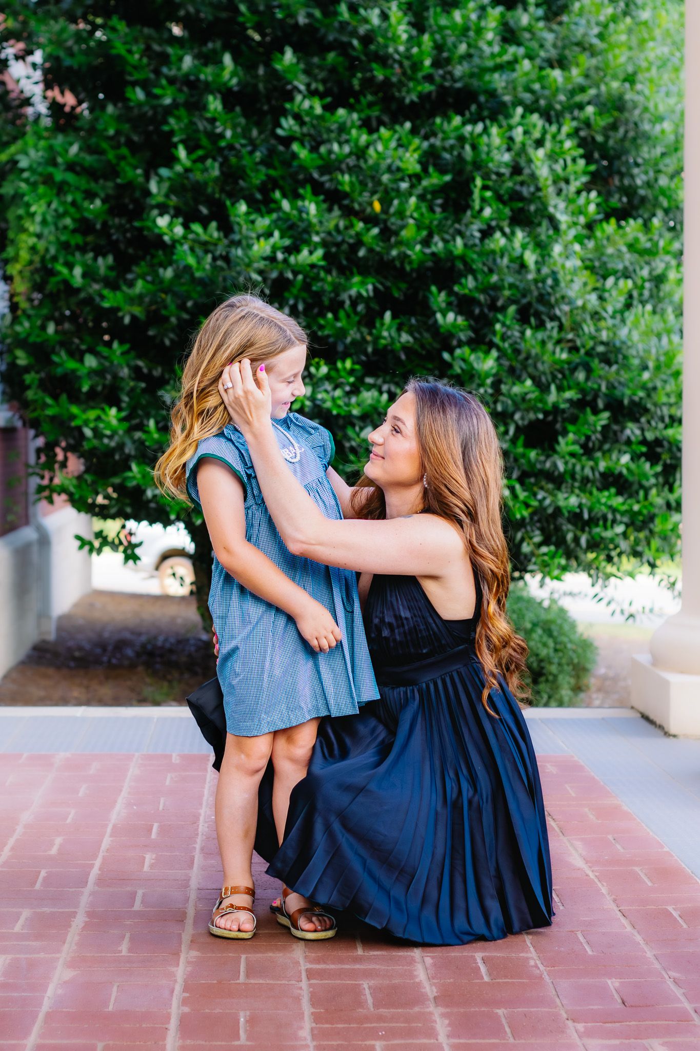 A mother crouches down to embrace her young daughter outdoors, both wearing blue dresses near lush green hedges.