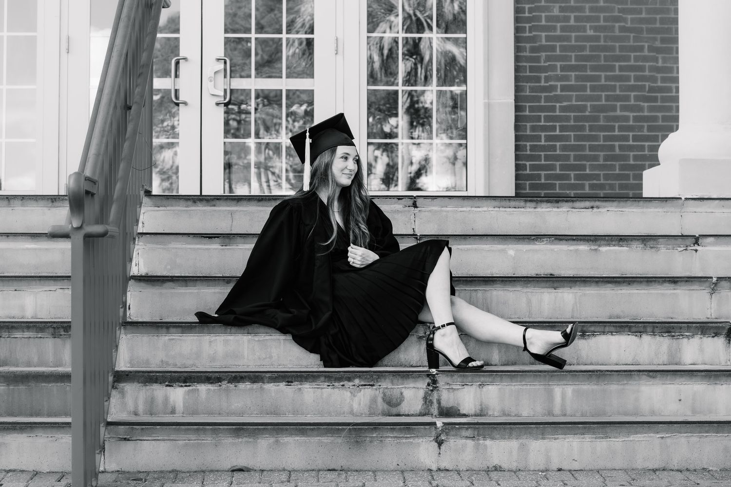 Young woman in graduation cap and gown sitting on university steps in black and white photo.