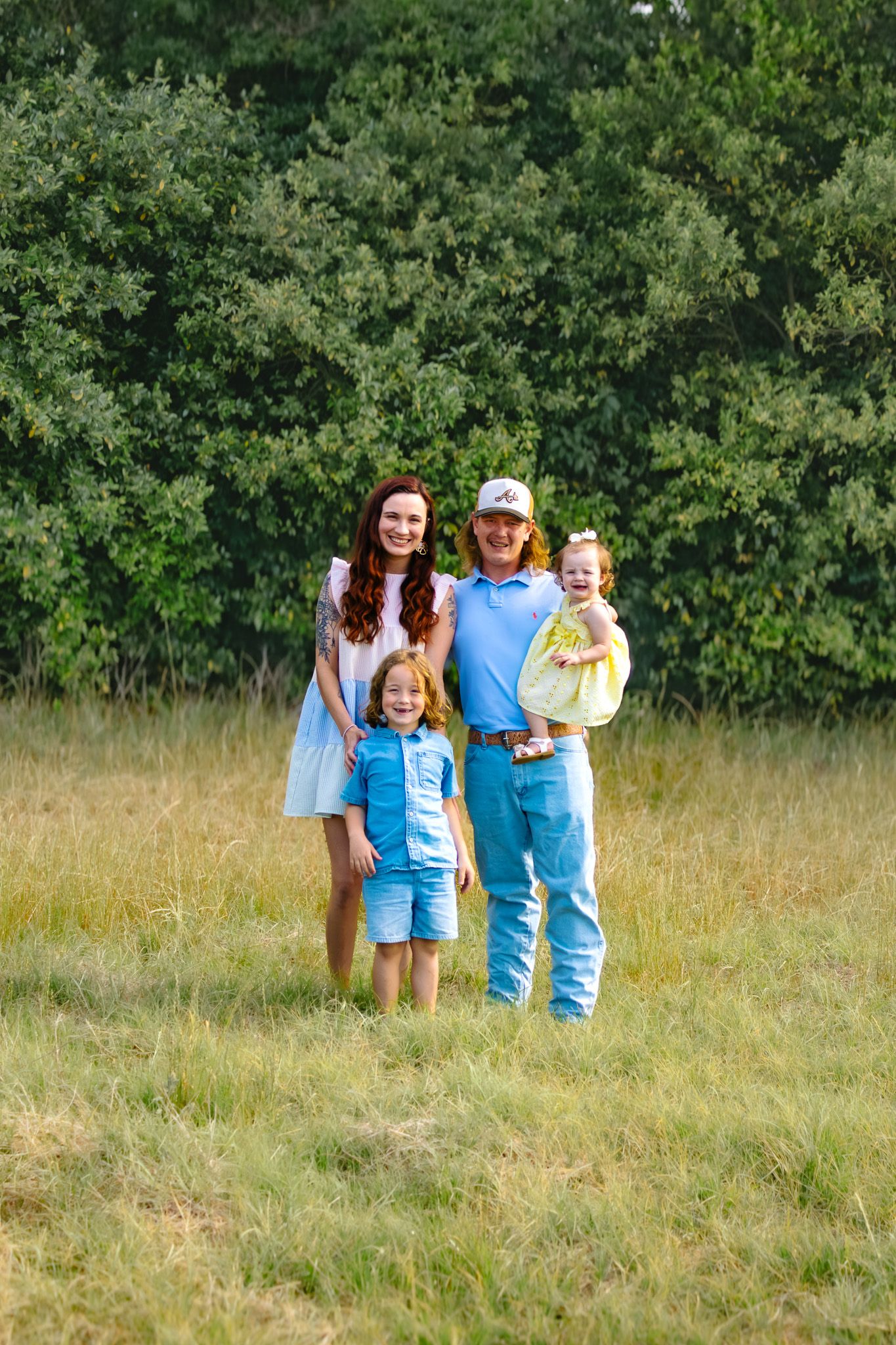 Happy family of four posing in a grassy field with lush green trees in the background on a sunny day.