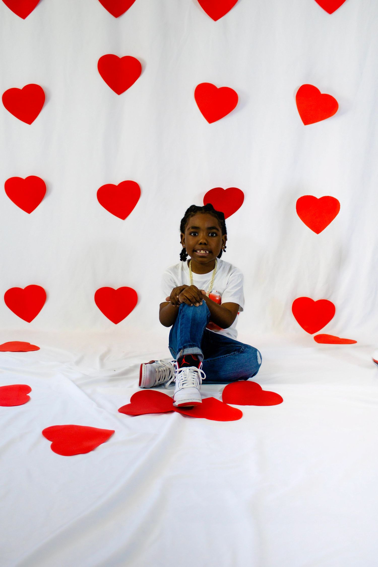 Young Black girl sits cross-legged on white backdrop with red hearts, wearing jeans and white tee, holding lollipop.