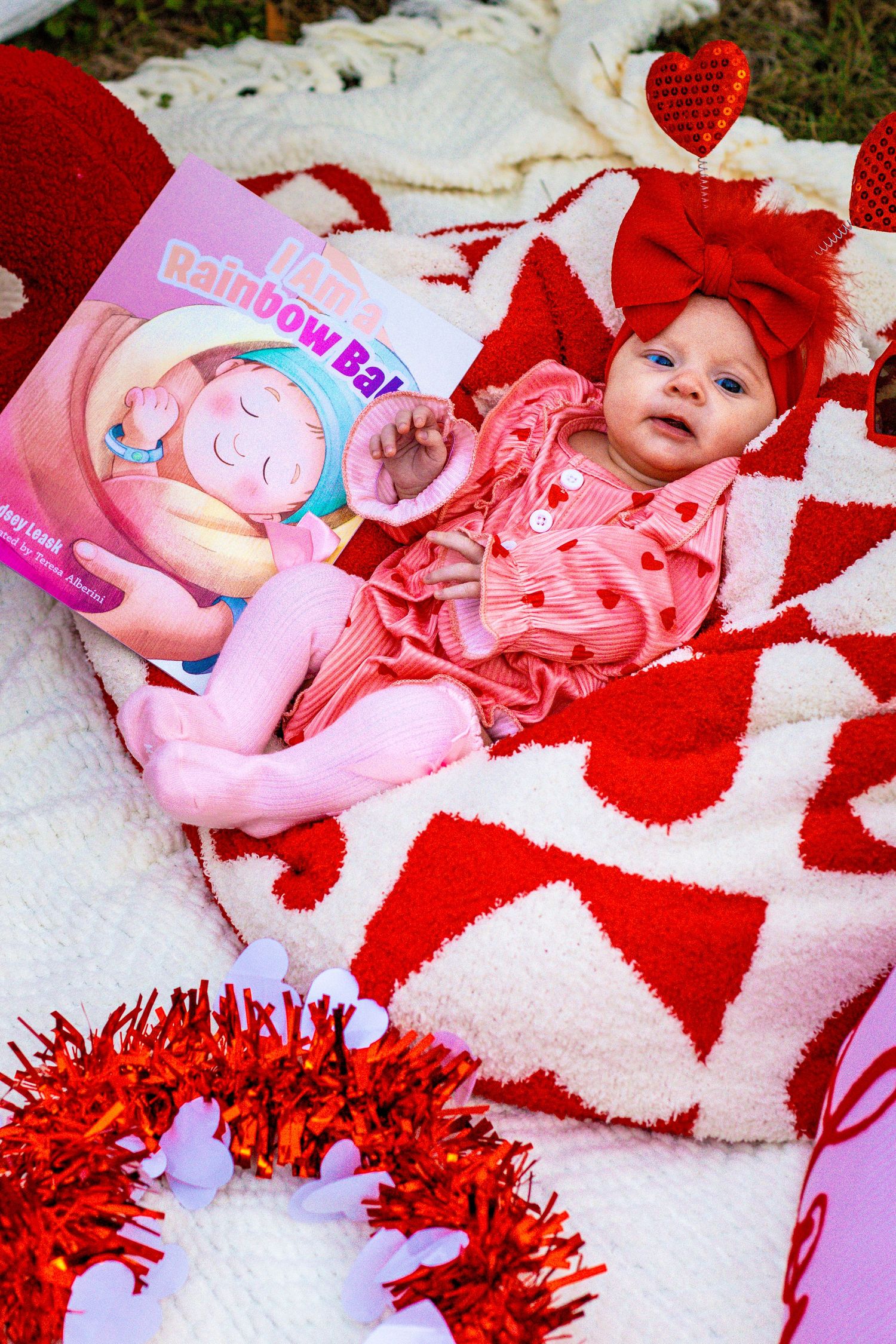 Newborn baby in pink outfit with red bow lies on heart blanket beside Rainbow Baby book and red tinsel.