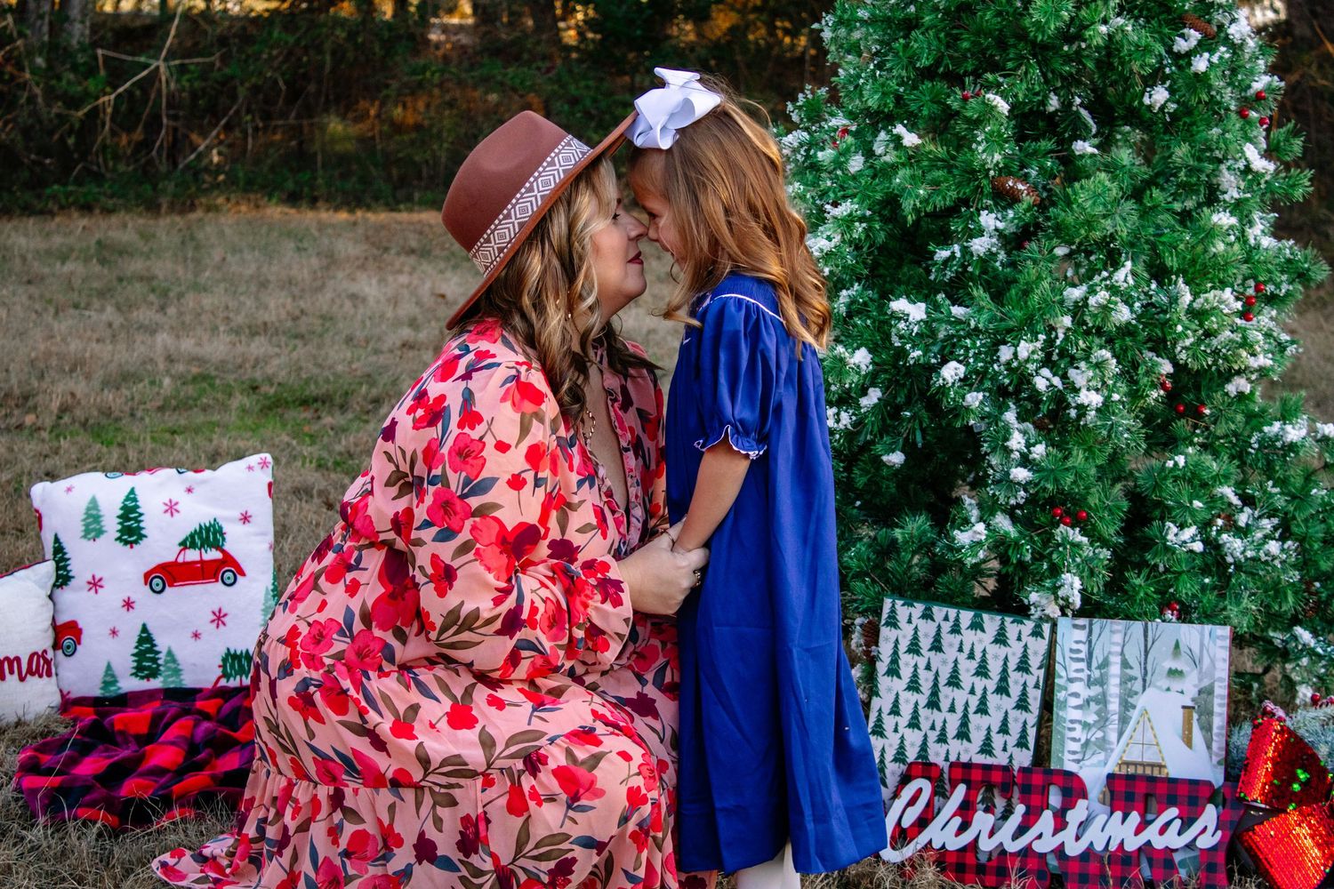 A mother and daughter share a tender nose-to-nose moment beside a Christmas tree outdoors.