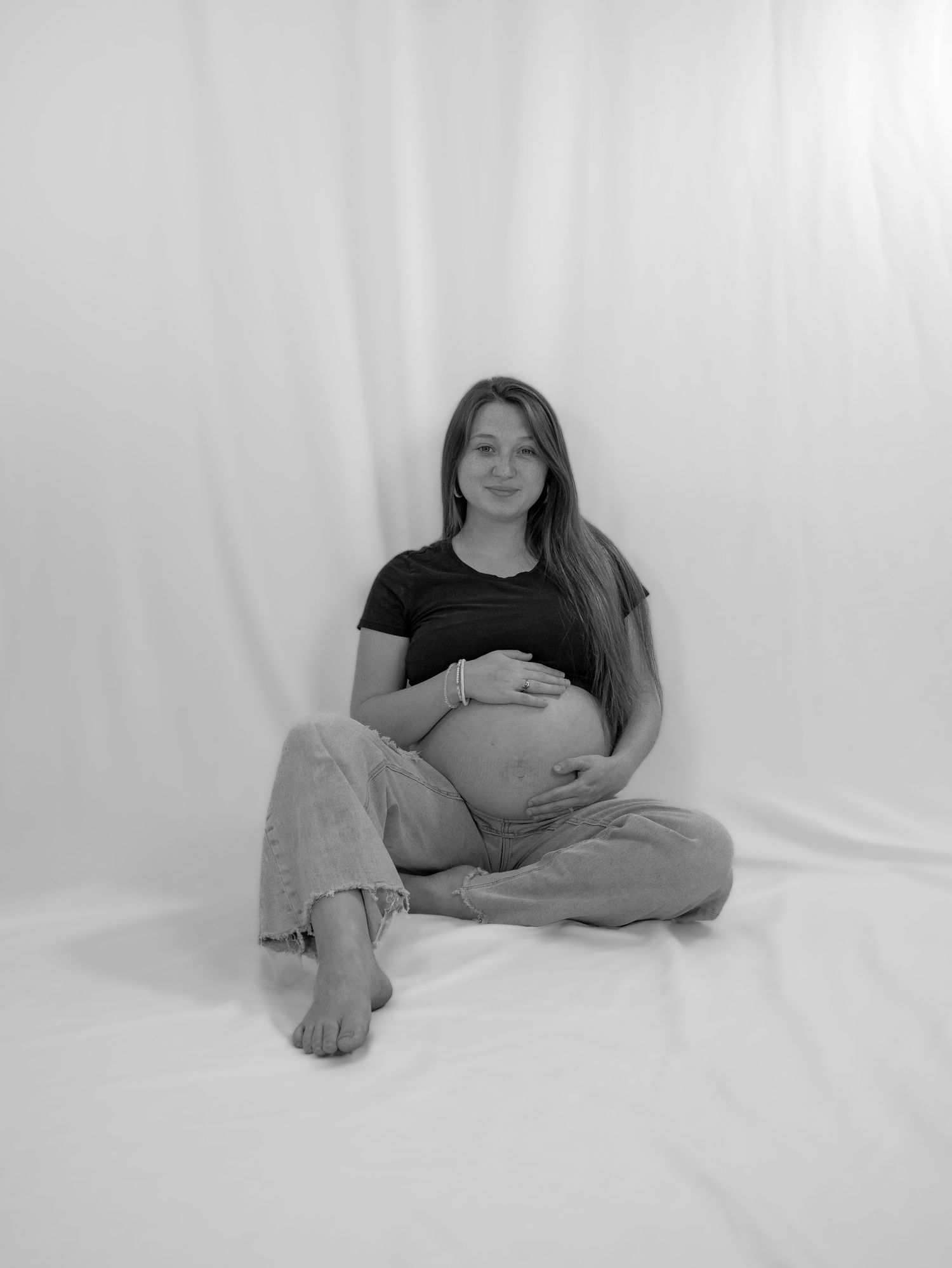 Black and white studio photo of pregnant woman sitting cross-legged on floor, cradling her bare baby bump.