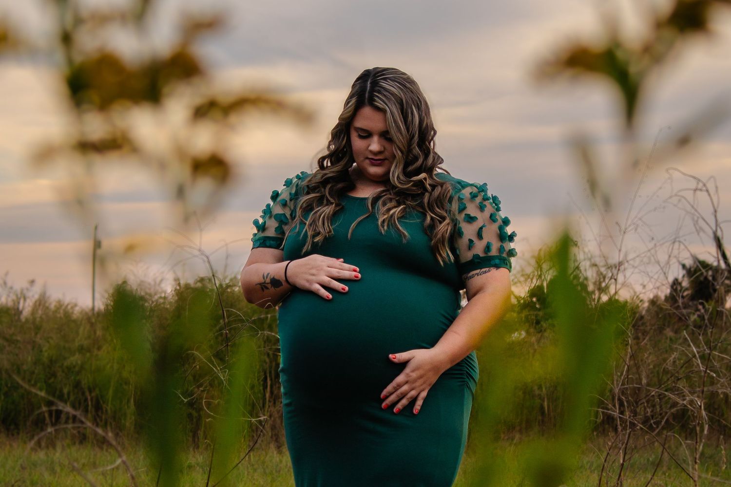 Pregnant woman in green dress cradling baby bump in golden field at sunset.